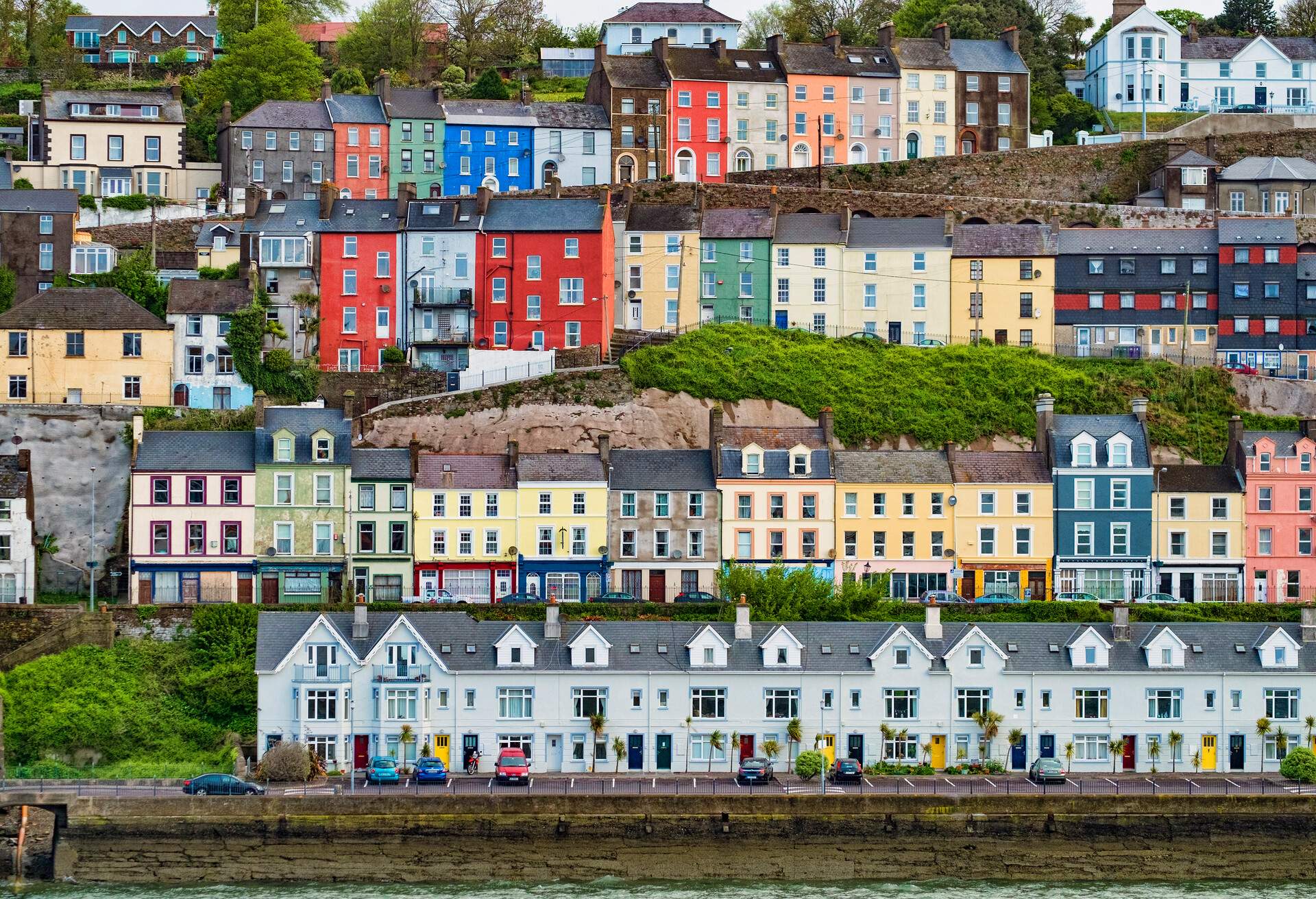 Horizontal image of Cobh, Ireland with colourful houses in rows on the hillside in town.  Houses can be seen in the bottom of the image at sea level and then proceeding up the hill to the top.  These are townhouses that are all attached in rows and are brightly coloured as well. The second from the bottom row of house are along the residential part of the Cobh high street.  Image taken in late summer from sea level.