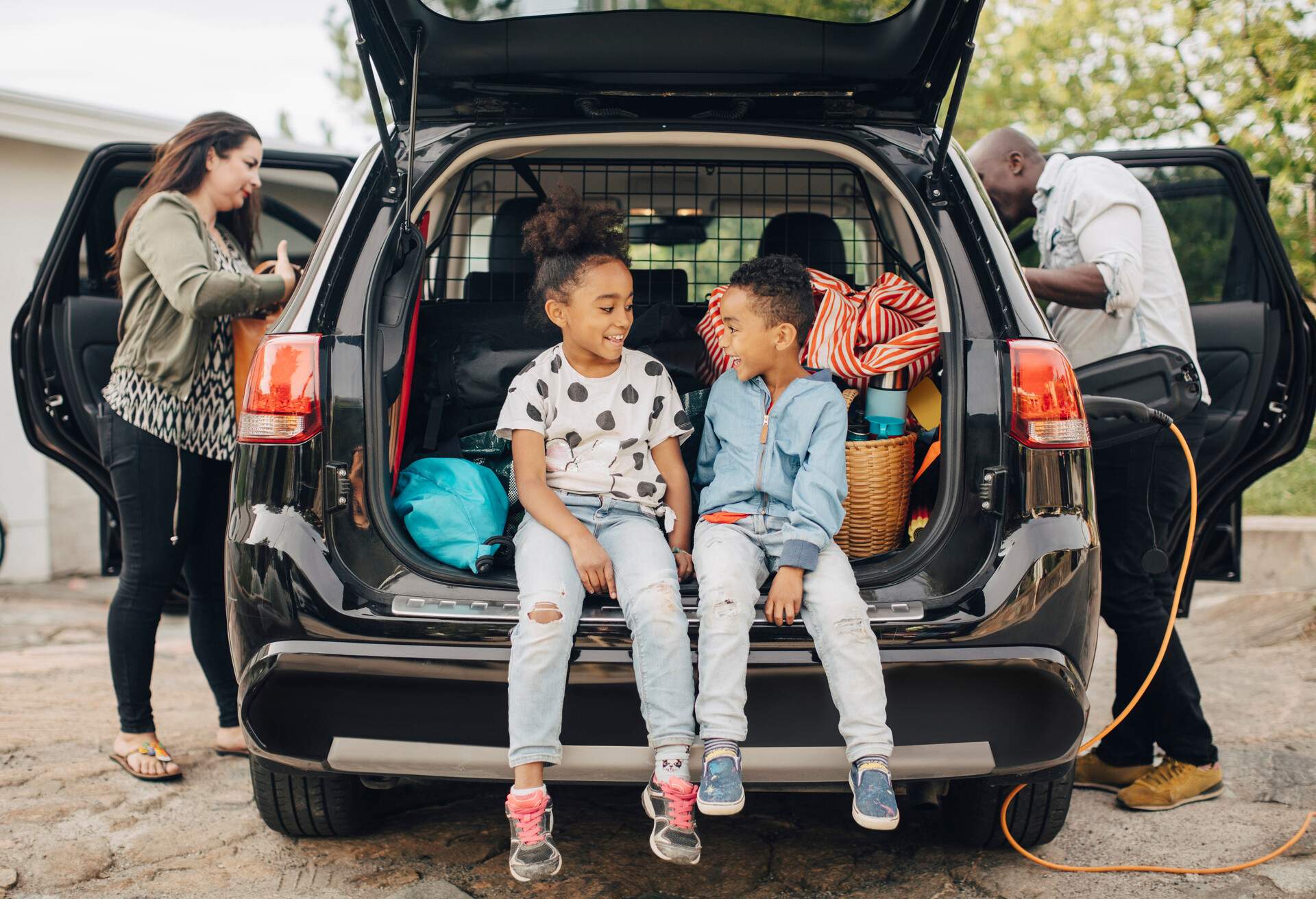 Smiling siblings sitting on car trunk with parents standing in front yard