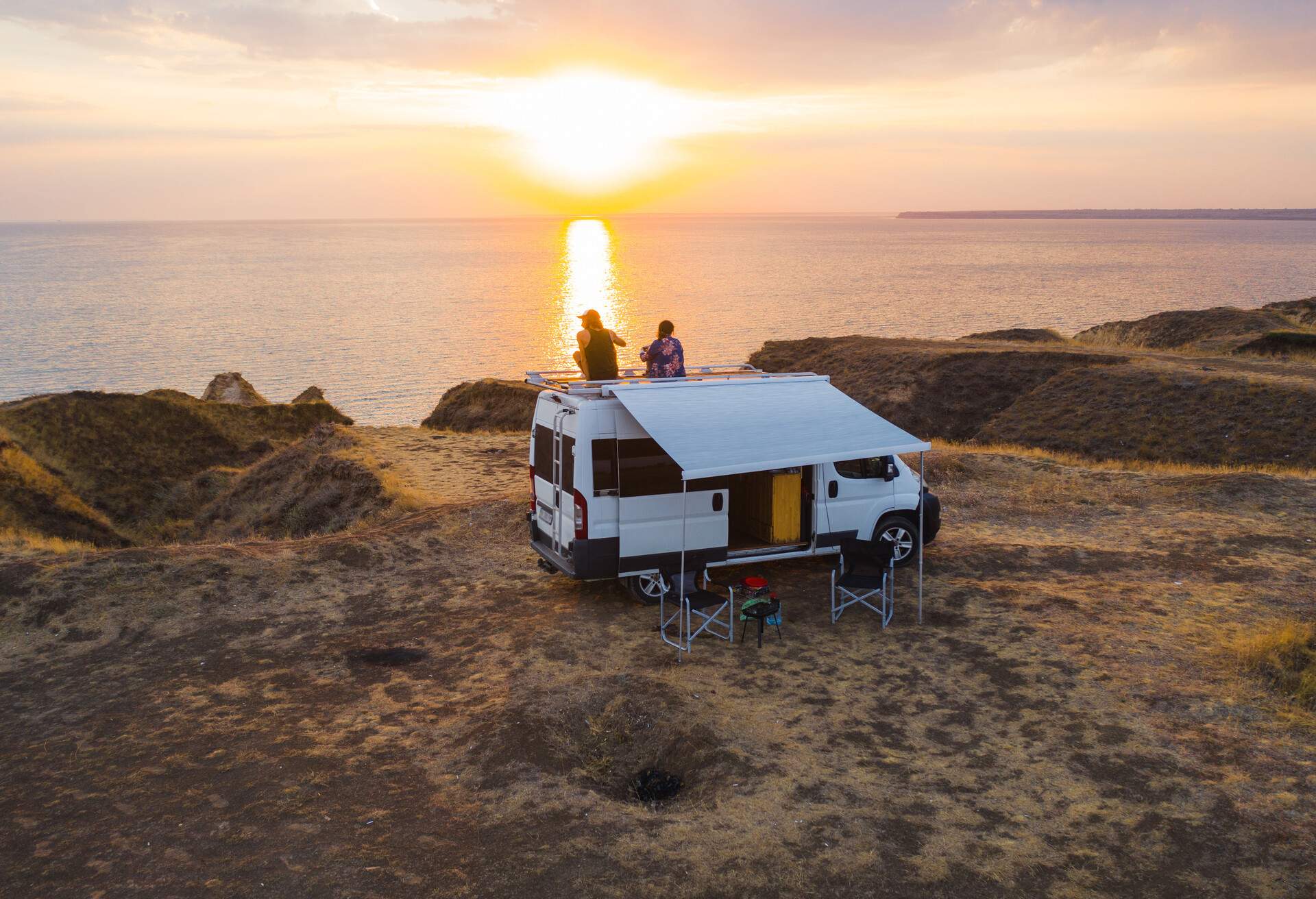 Aerial view of a couple on the roof of camper van on seaside at sunset.