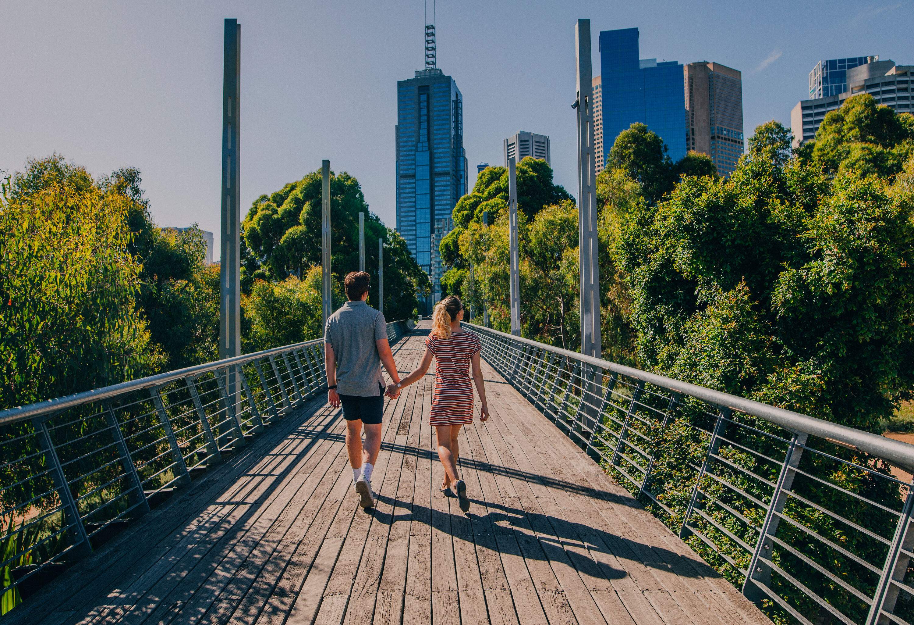 A lovely couple holds hands while walking on a wooden pedestrian bridge between the lush trees heading towards the tall buildings.