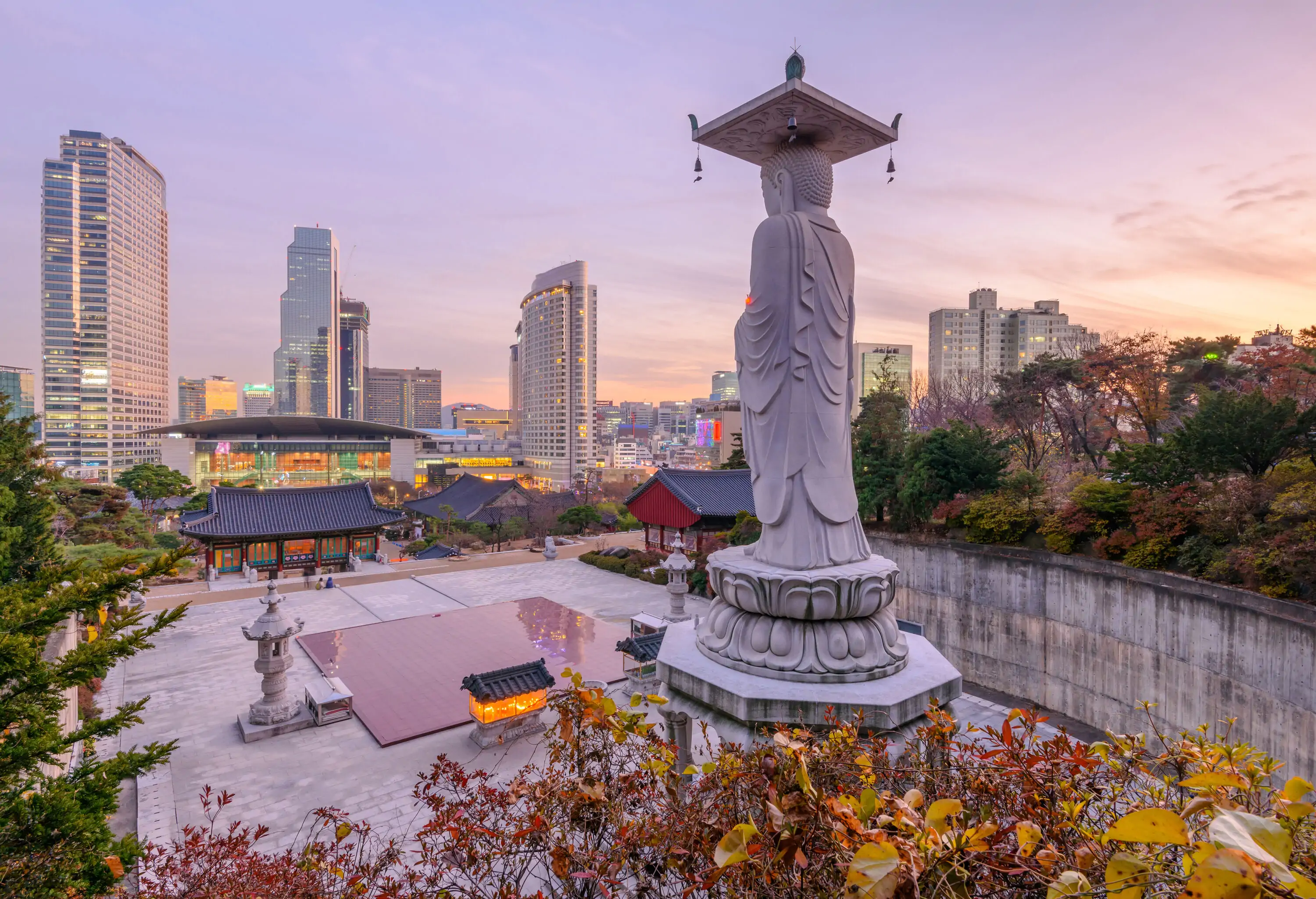 An enormous stone statue stands in a cobbled square surrounded by trees facing a Buddhist temple against the skyline.