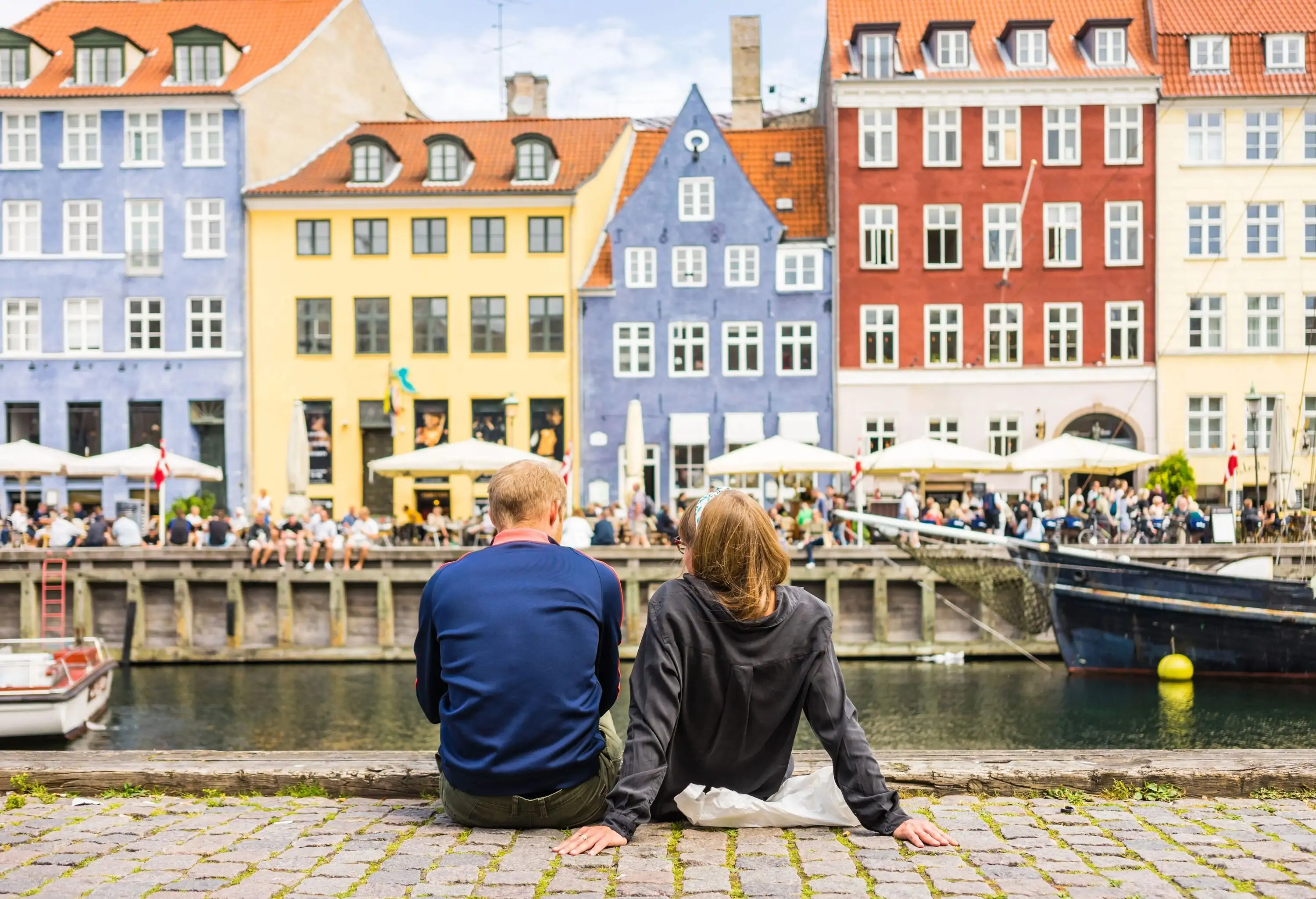 Two individuals sit on the promenade by the canal overlooking the pedestrians on the street lined with colourful and classic buildings.