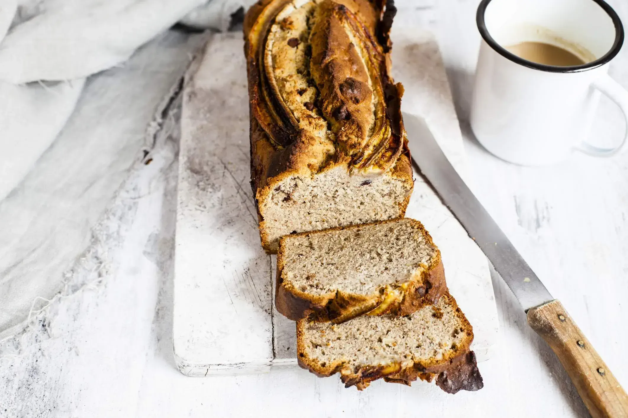 sliced banana bread loaf on a white wooden board with a knife and mug of coffee