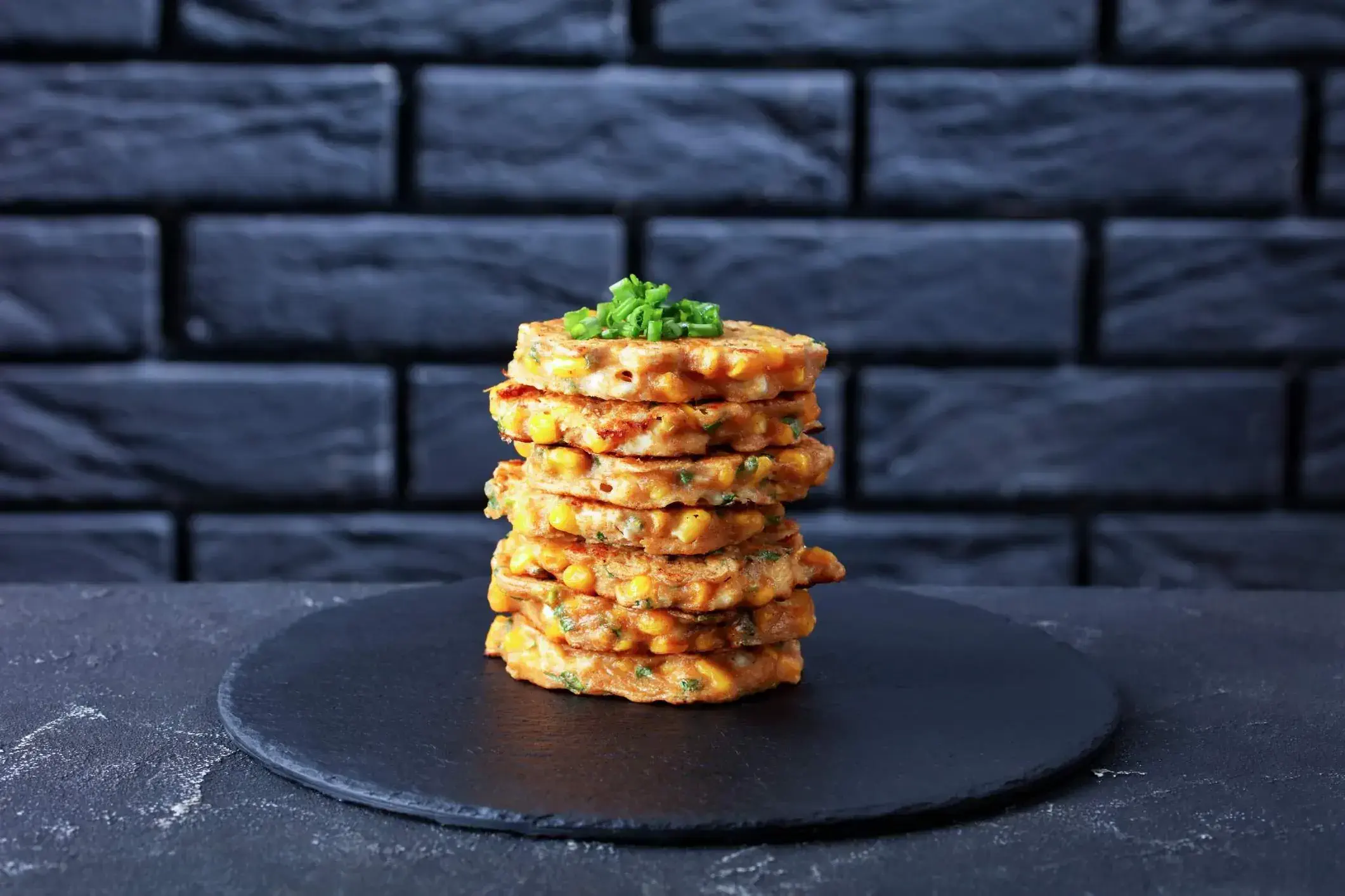 Black slate plate with stack of sweetcorn fritters in front of black brick wall