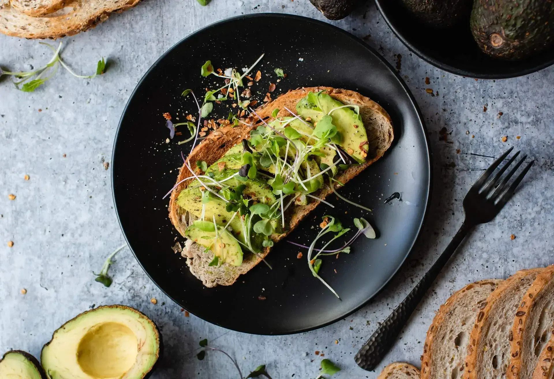 Black plate with a slice of avocado on toast and black fork