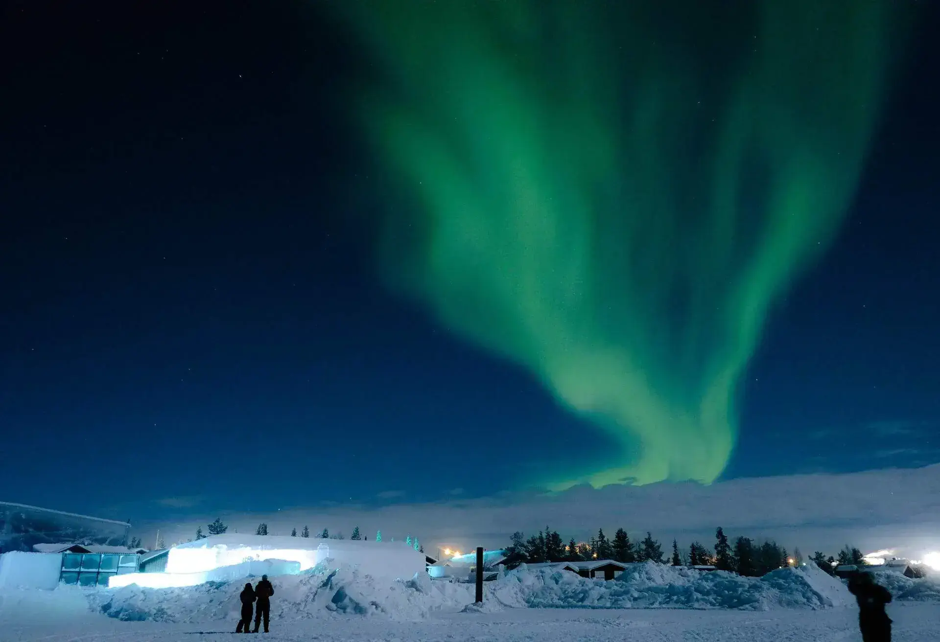 Silhouette of two people in a snowy resort looking at the huge sky with Northern Lights