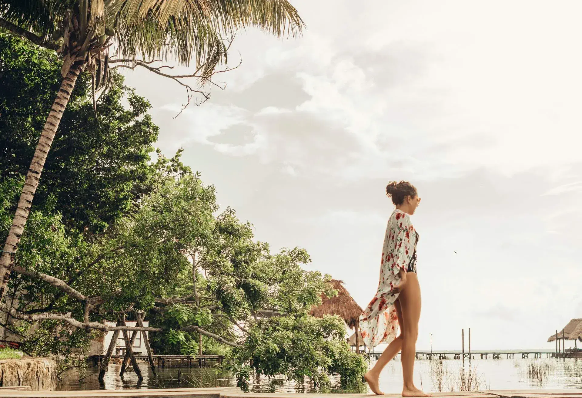 Woman wearing a floral kaftan walking by a lagoon surrounded bz tropical shrubbery on an overcast day