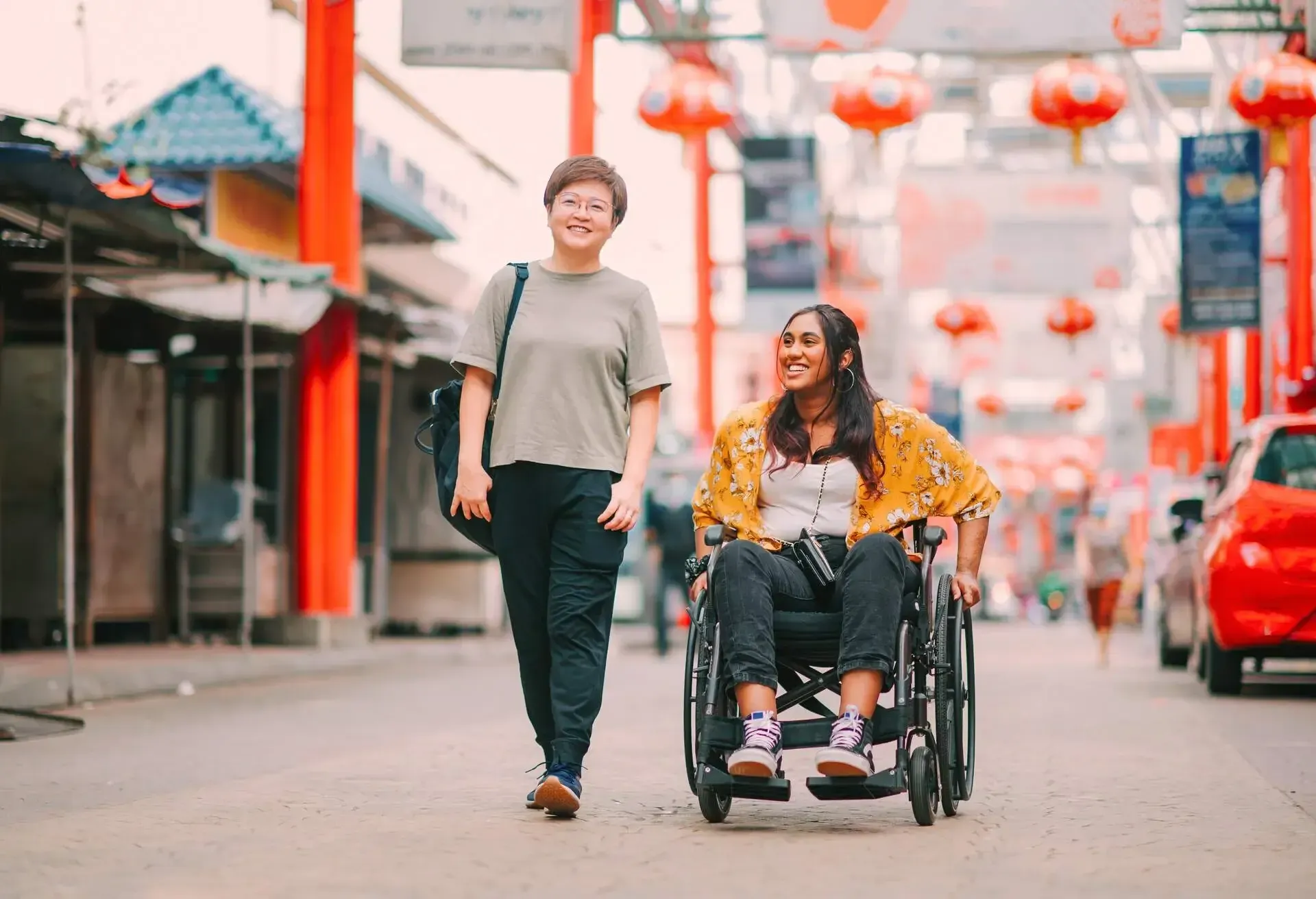 Two female friends in Asian city smiling and walking on a pedestrian street, one in a wheelchair