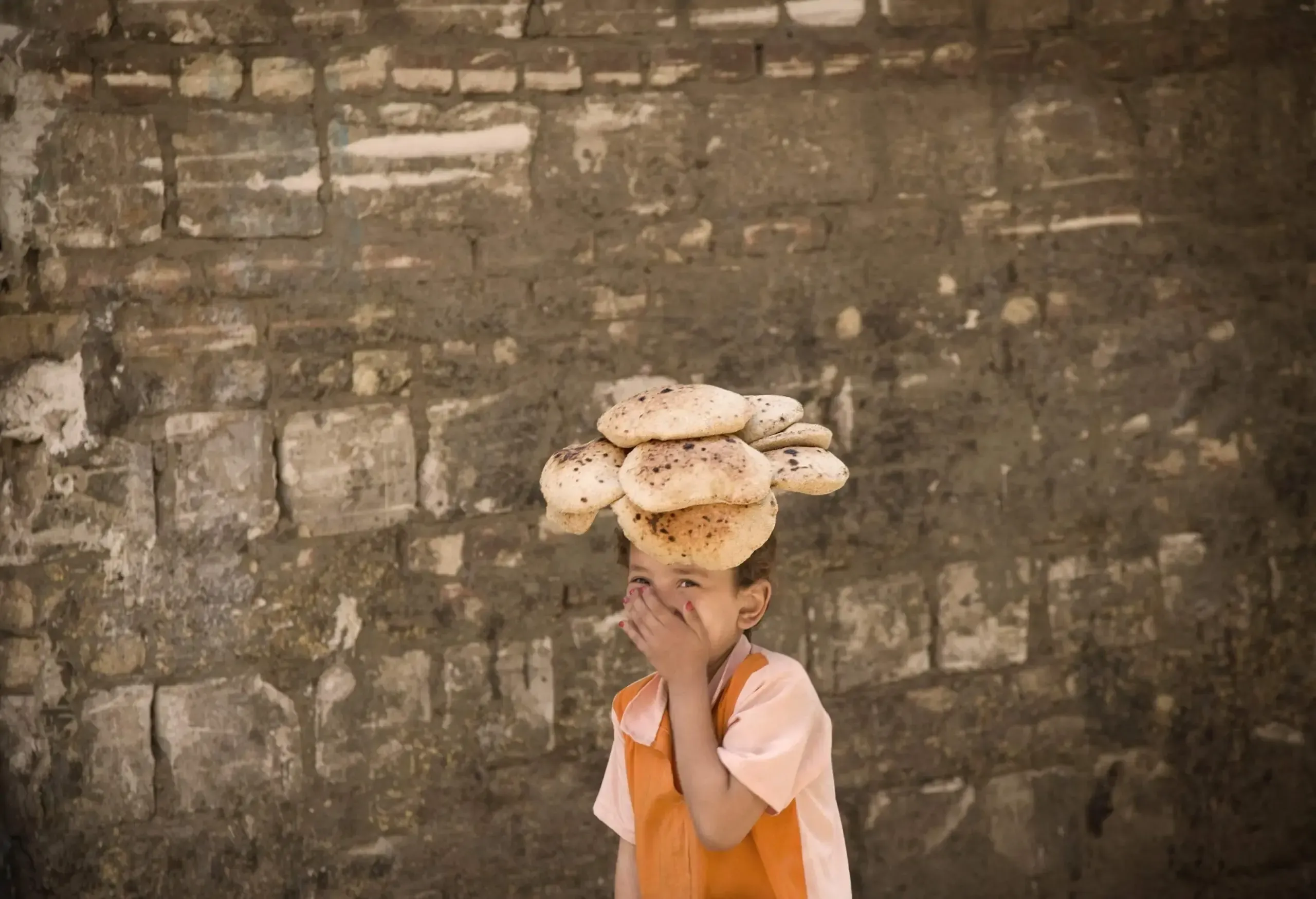 Laughing child covering his mouth carrying bread on his head by a wall