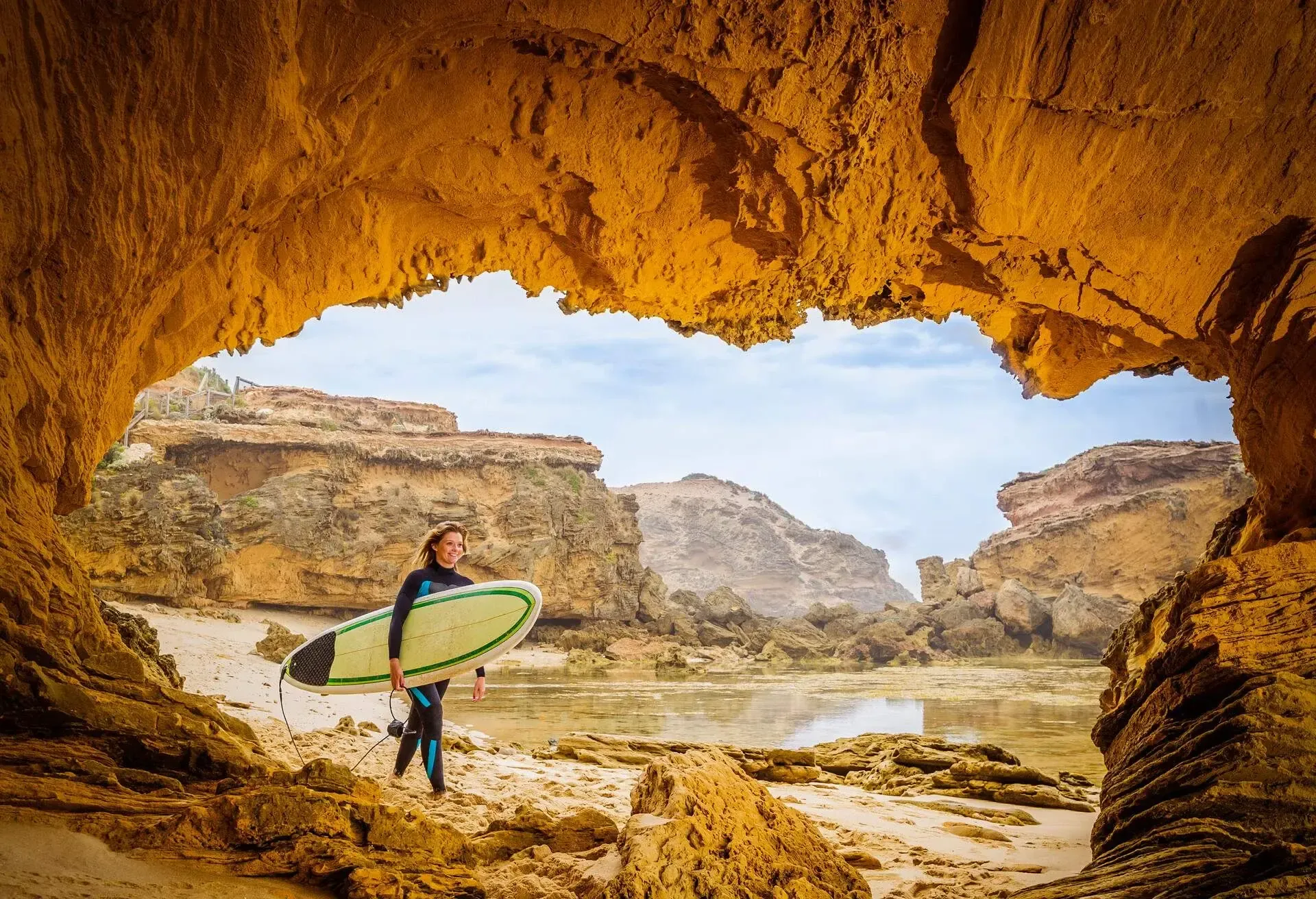 View from a beach cave of a female surfer walking on a rocky beach holding a surfboard