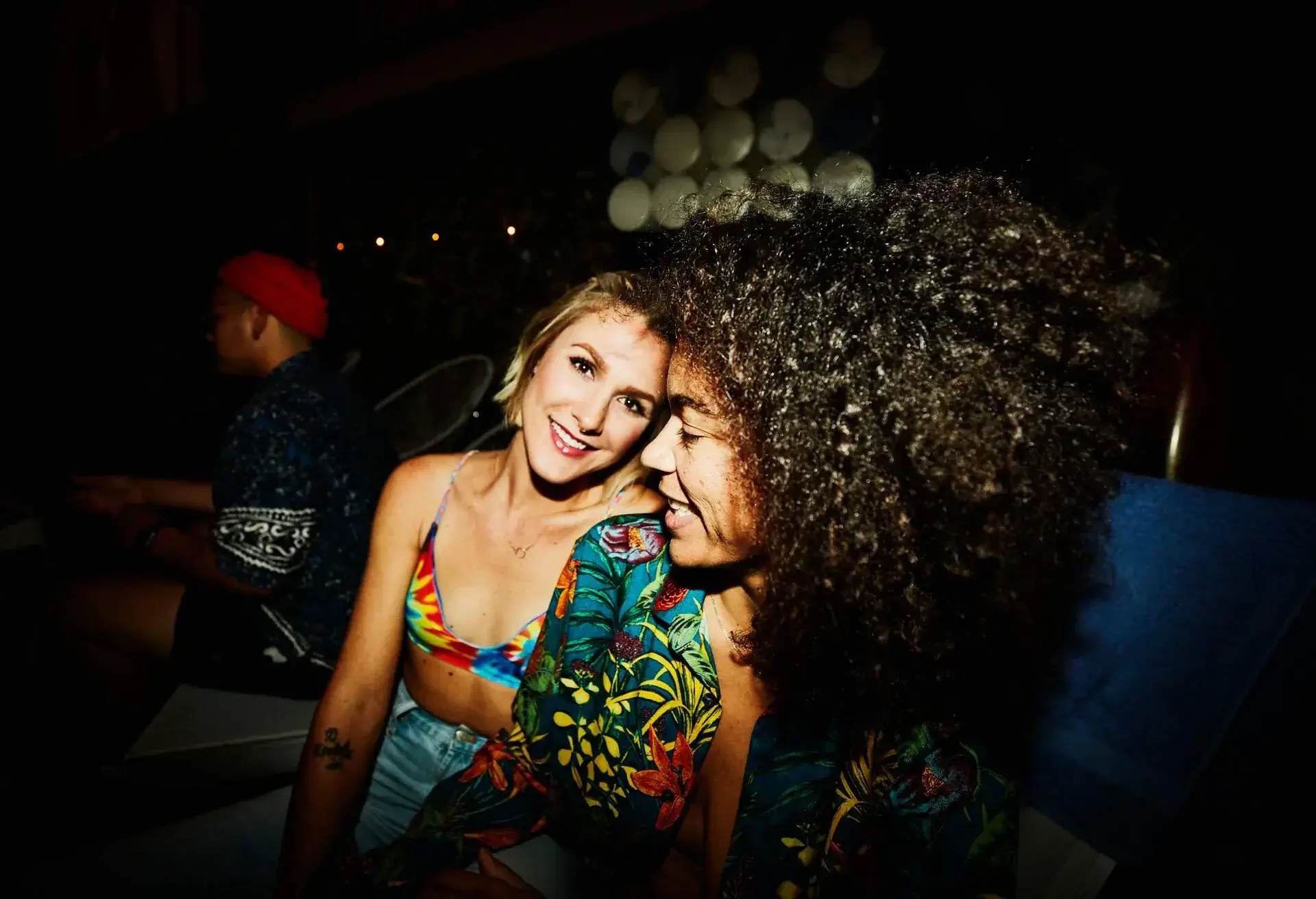Two friends pose closely together, wearing vibrant outfits with tropical patterns, in a dimly lit social setting.