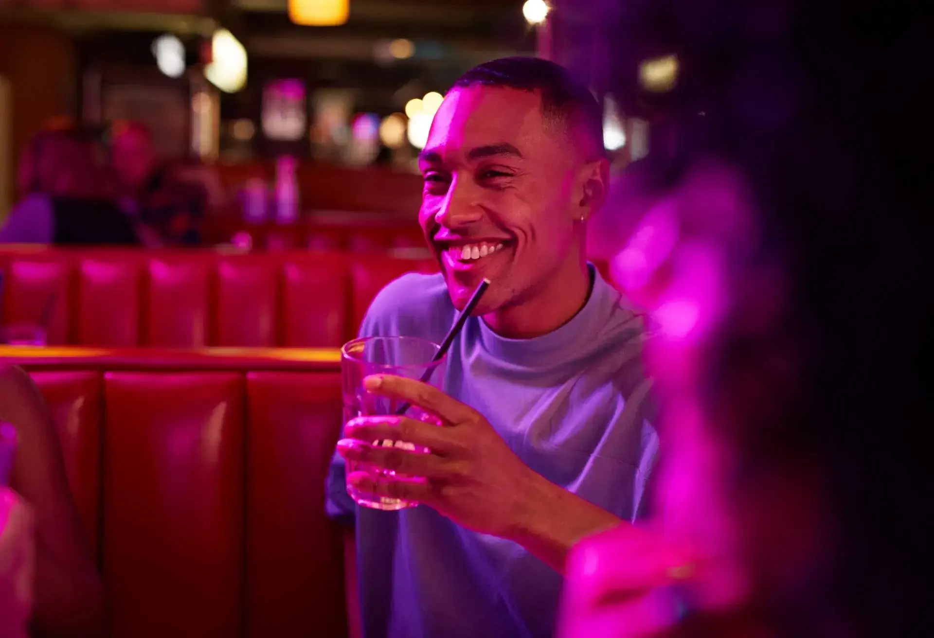 A person wearing a light blue shirt holds a glass with a straw in a vibrant, dimly lit setting with red booths.