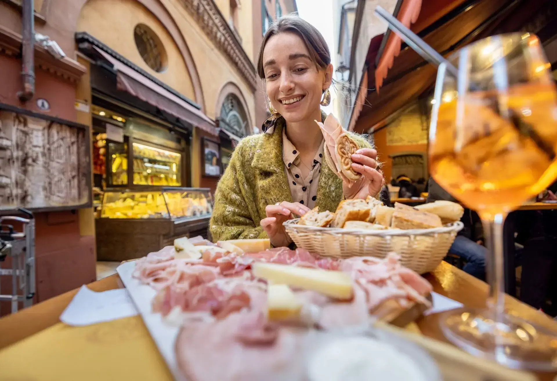 A woman dining outdoors served with sliced meats, cheese, a basket of bread, and a glass of drink.