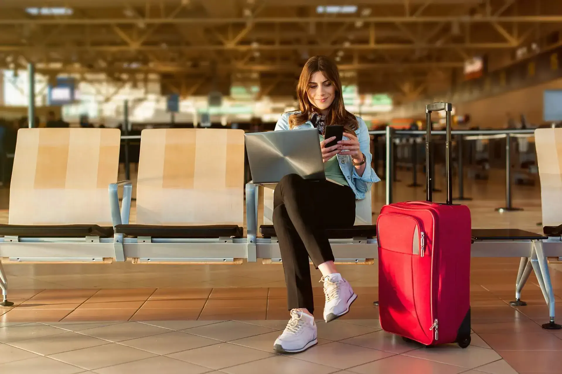 A woman using her laptop and phone while sitting next to her pink suitcase at an airport terminal.