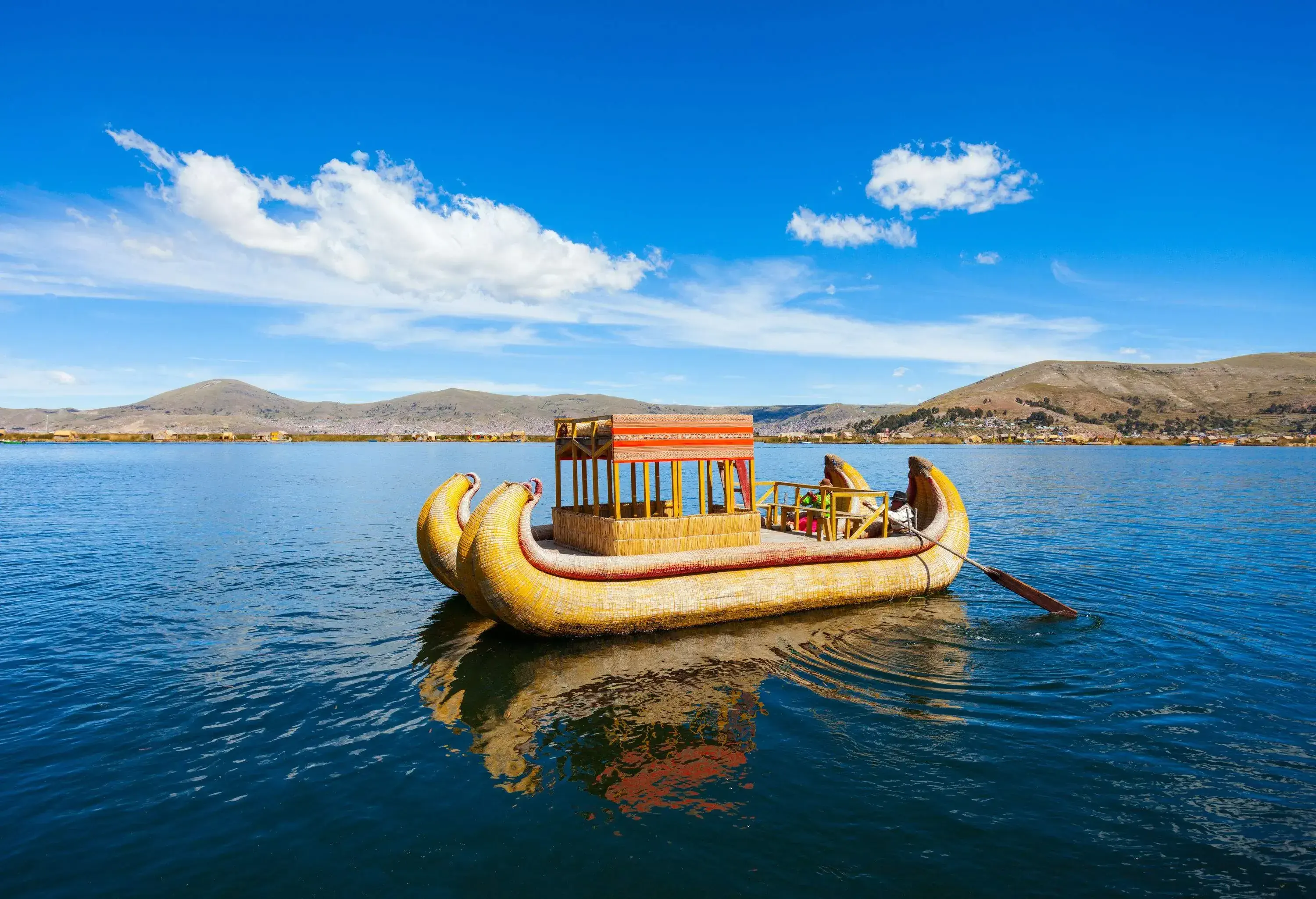 Two people ride a yellow totora boat with a square shed and use a paddle to navigate the lake.