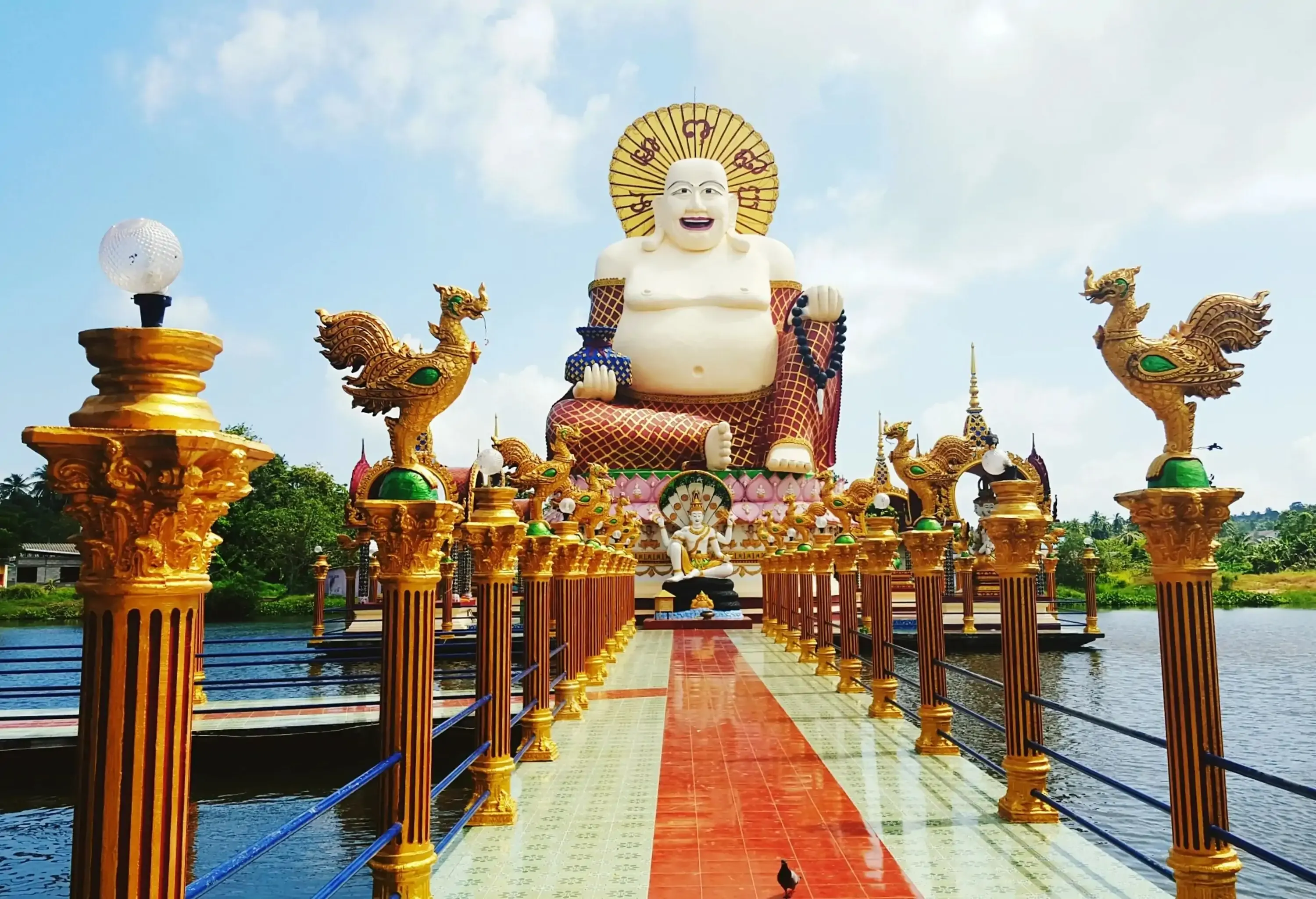 A large Chinese-style laughing Buddha at the end of a tiled walkway that spans a lake and is flanked by columns topped with gilded rooster figurines.