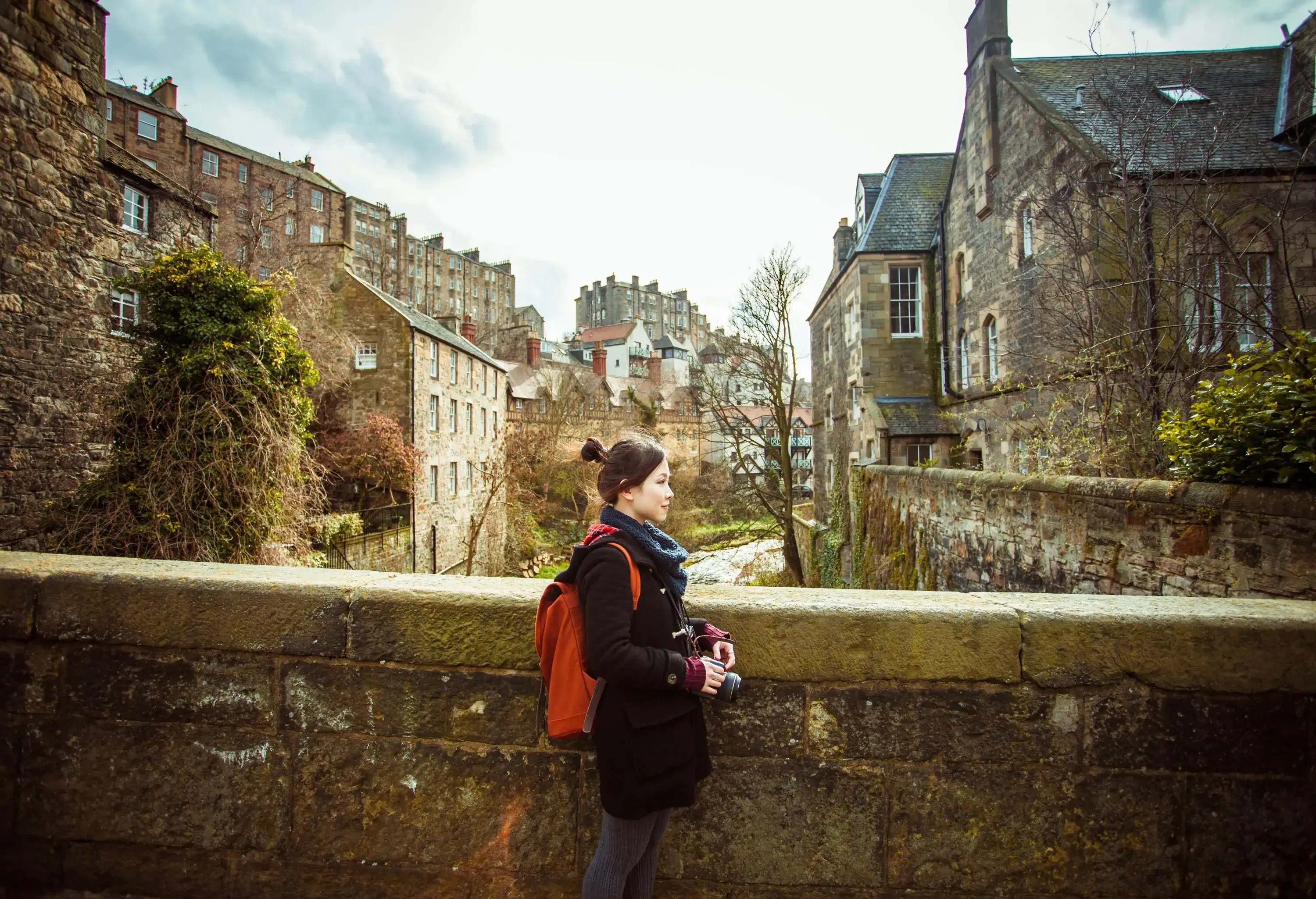 A person dressed for the cold with a backpack and a camera stands on an old bridge next to a brook surrounded by stone homes.