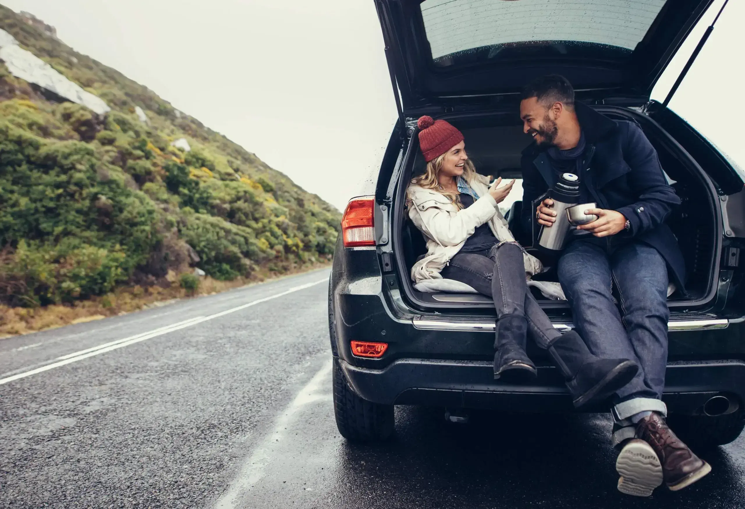 A joyful couple sits at the back of their parked car on the side of the road, exuding happiness and relaxation during their journey.