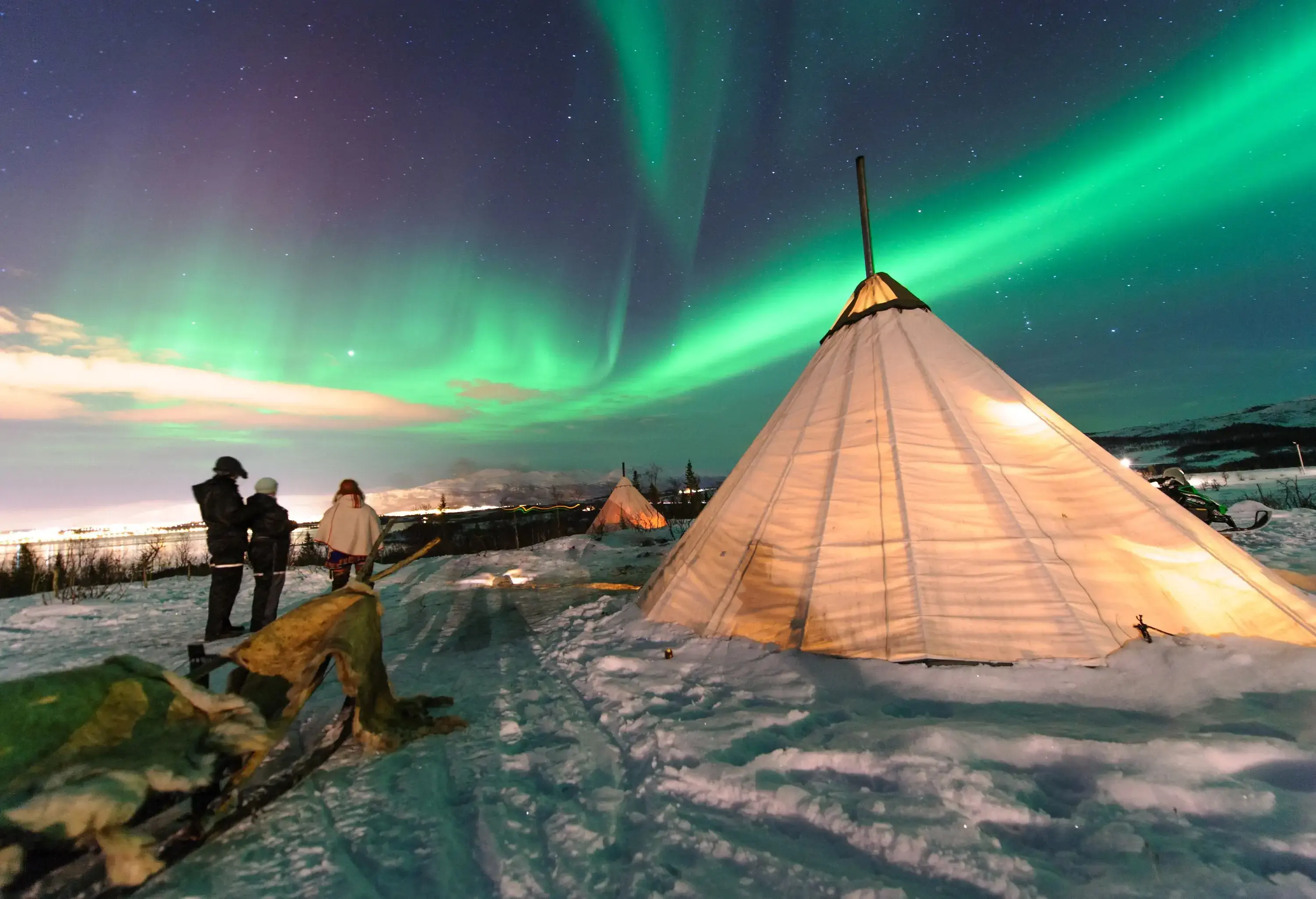 Three people standing on a snowfield strewn with teepee tents as the northern lights dance across the sky.