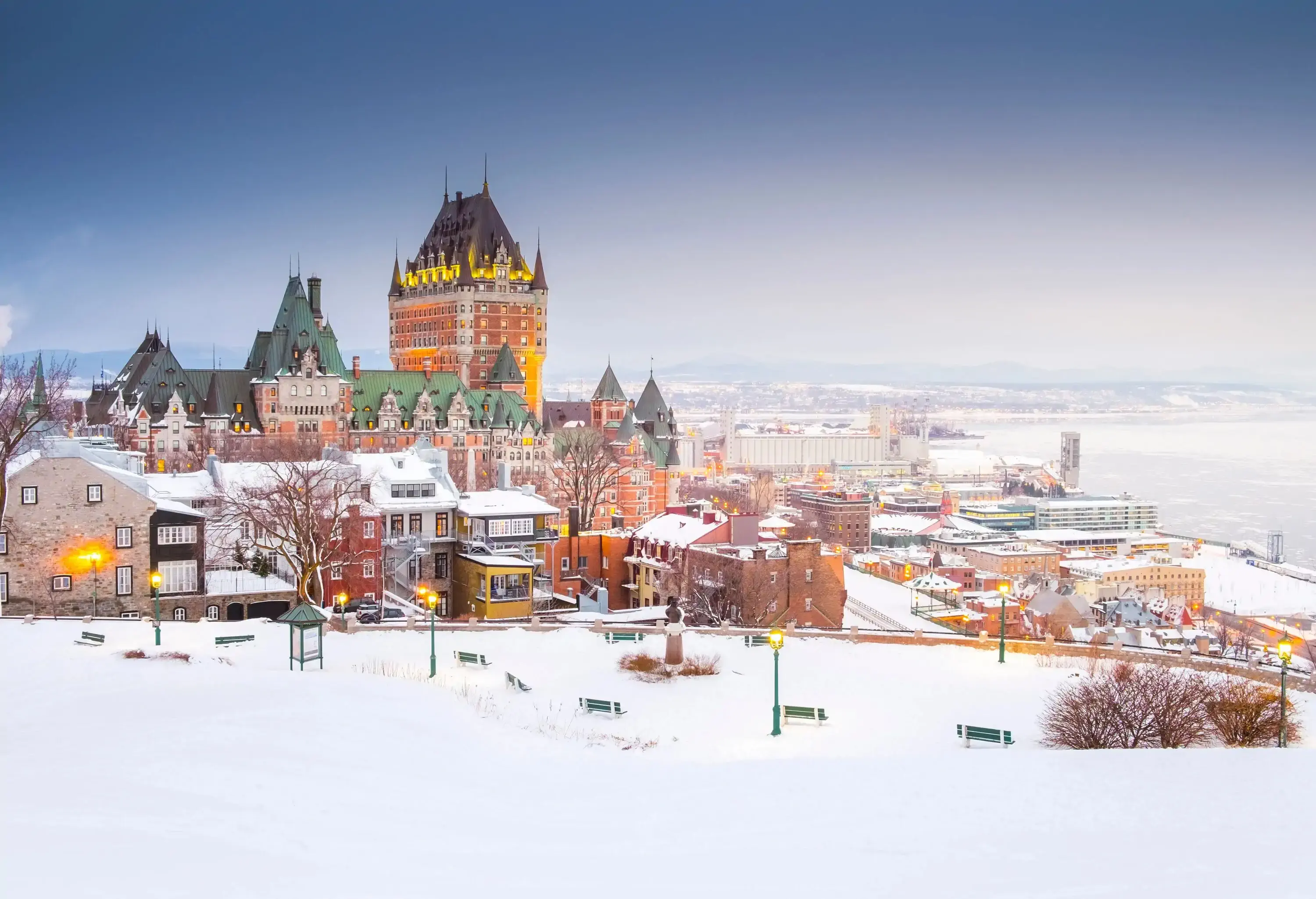 The Fairmont Le Château Frontenac hotel is situated in a snow-covered city near a frozen river.