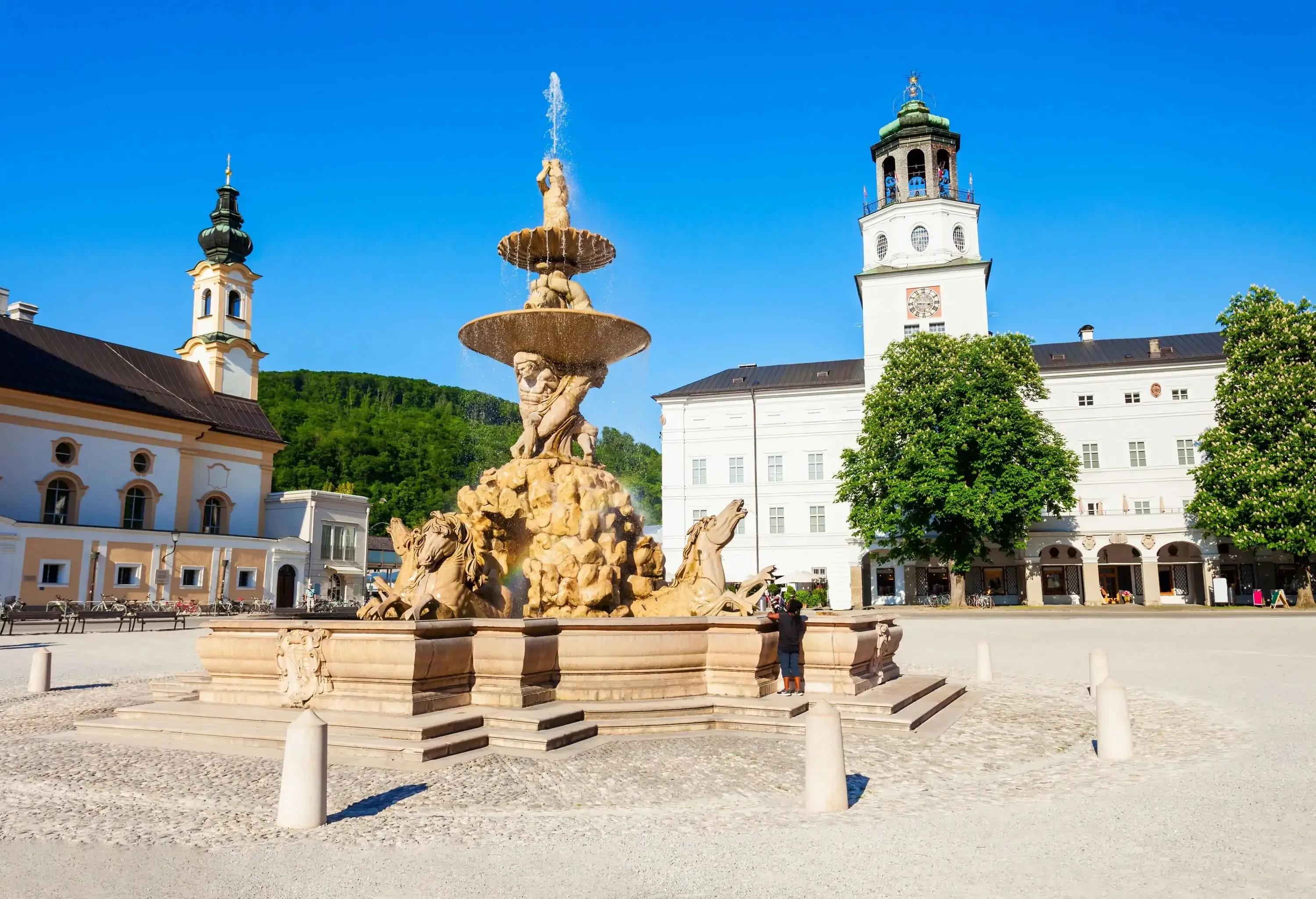 A public square with a sculptured fountain in the centre surrounded by buildings with a clock tower and towering spire.