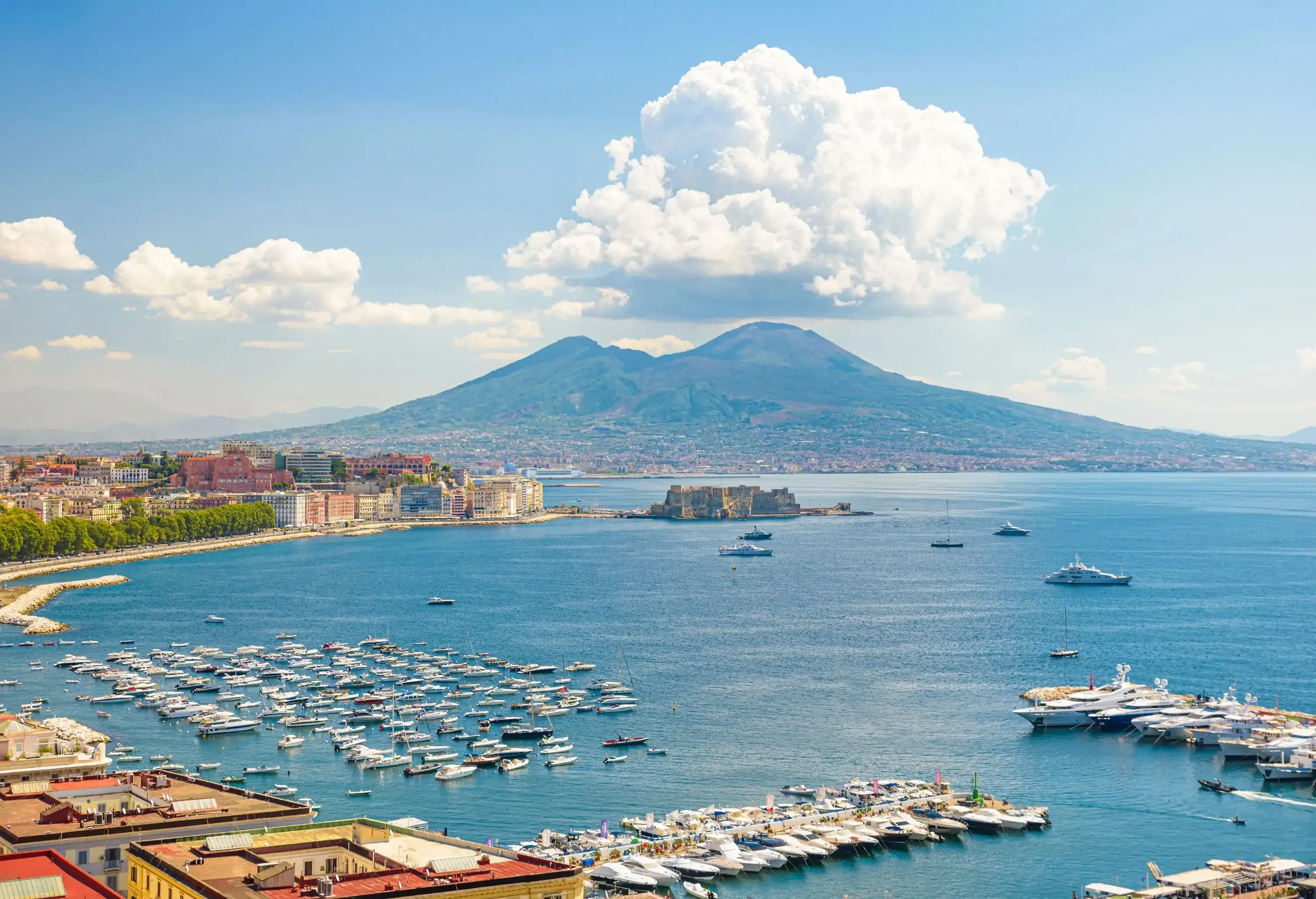 The bustling harbour sits along the sparkling blue sea, as a charming waterfront town buzzes with life against the backdrop of a majestic volcano in the distance.