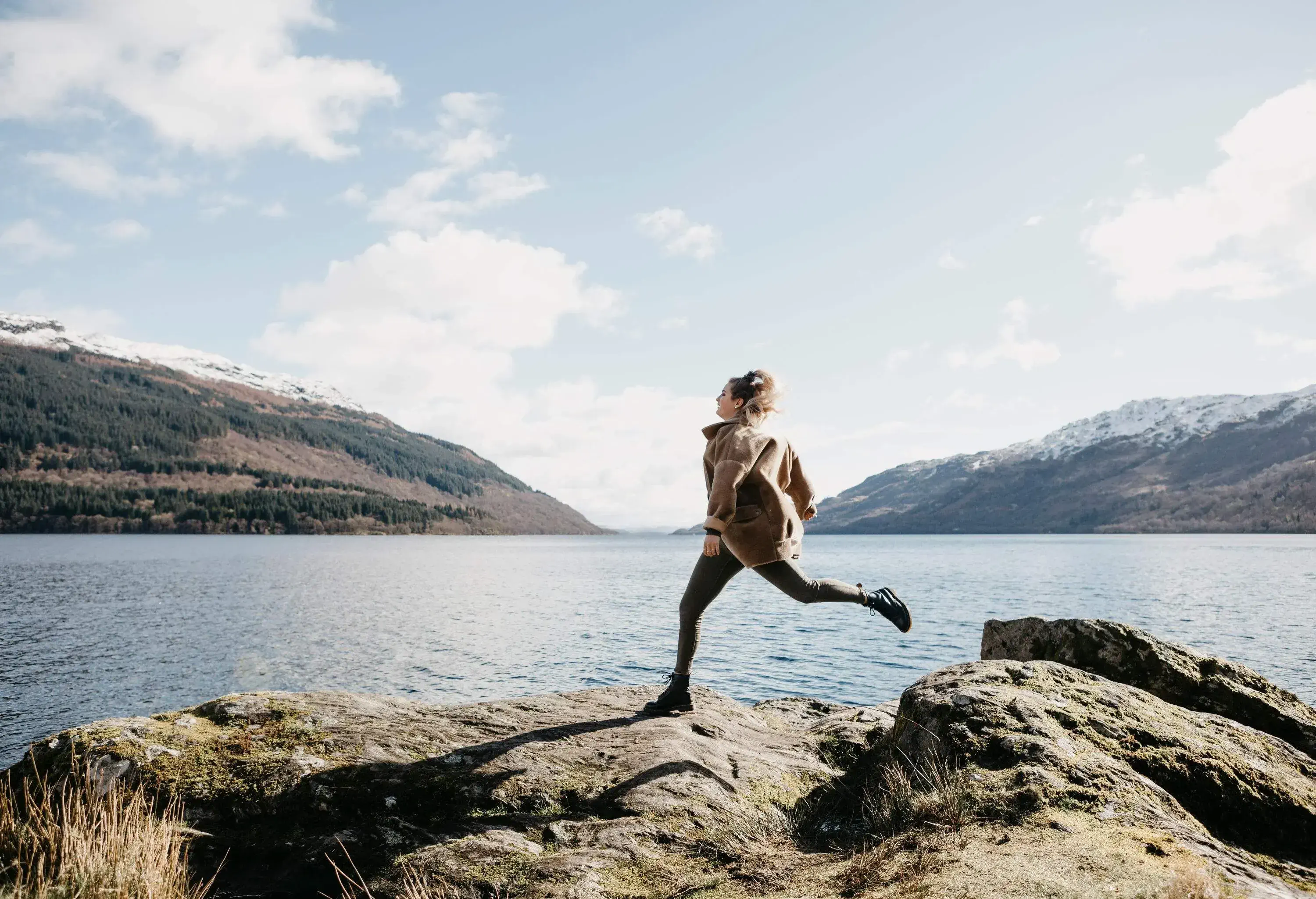 Woman stepping over rocks wearing a winter jacket by a lake on a sunny winter day