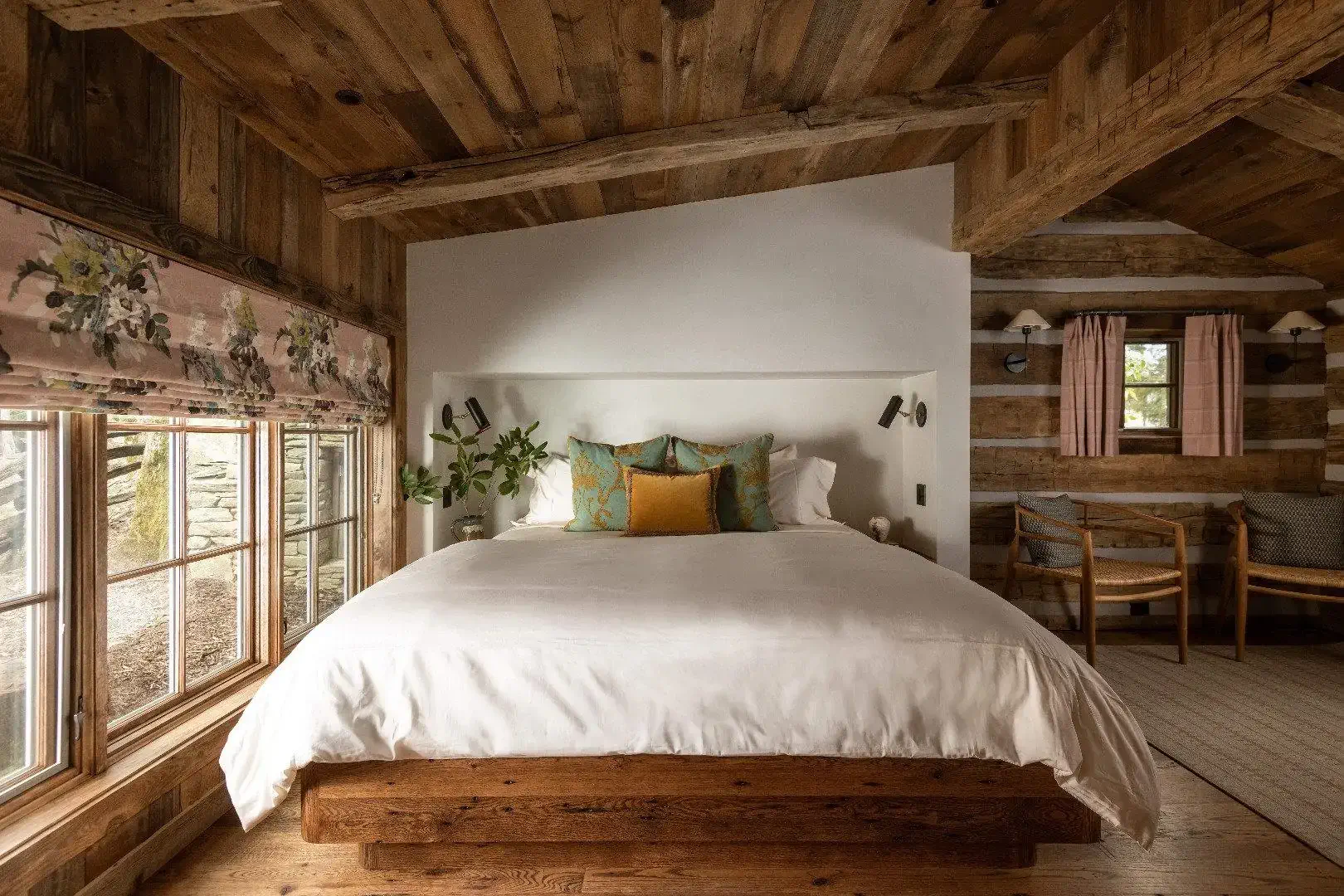 Cozy rustic bedroom with wood beams, large bed, patterned curtains, and wall lamps.