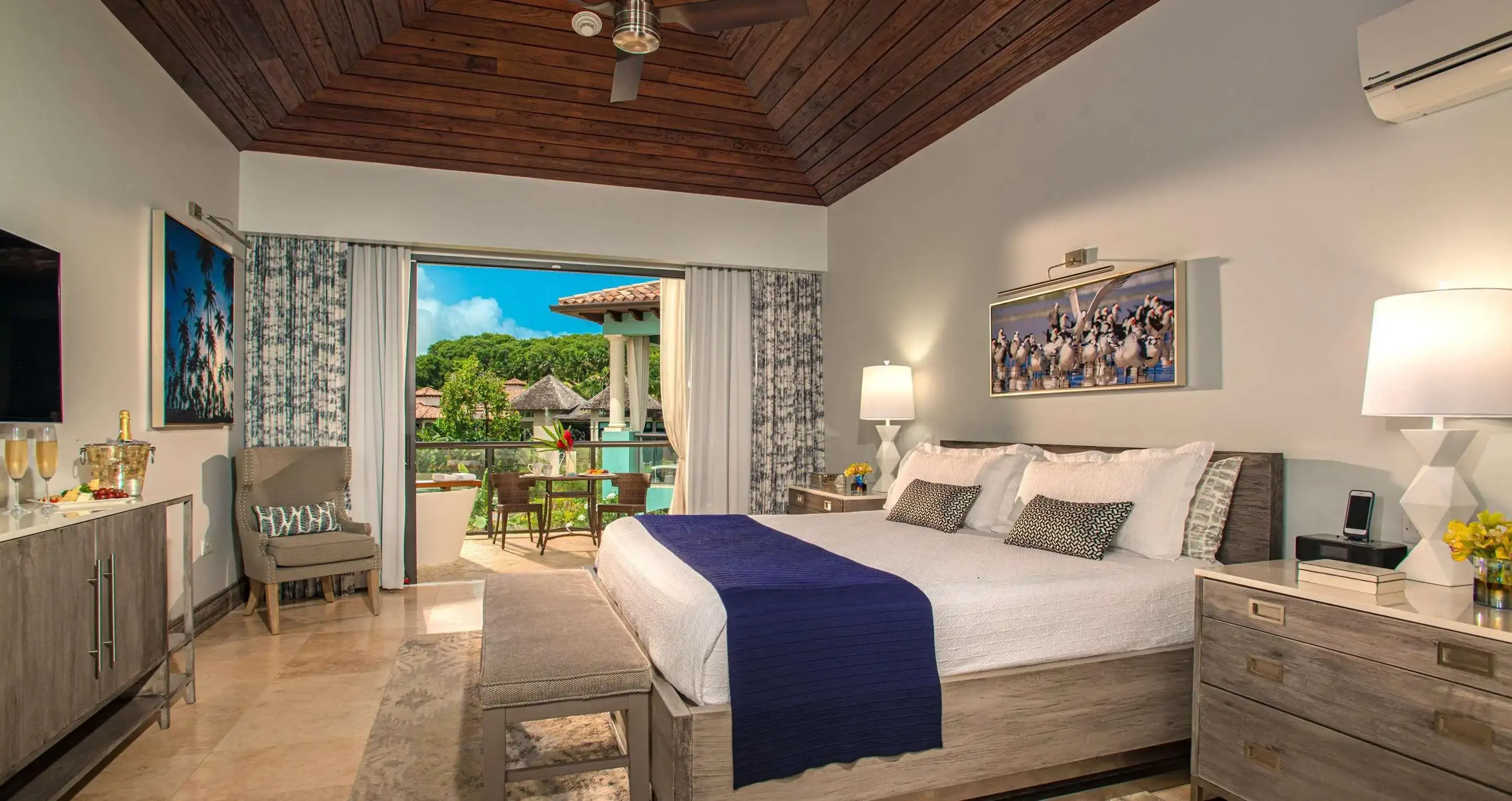 Luxurious hotel room with a king-size bed, balcony view, modern furnishings, and tropical decor.