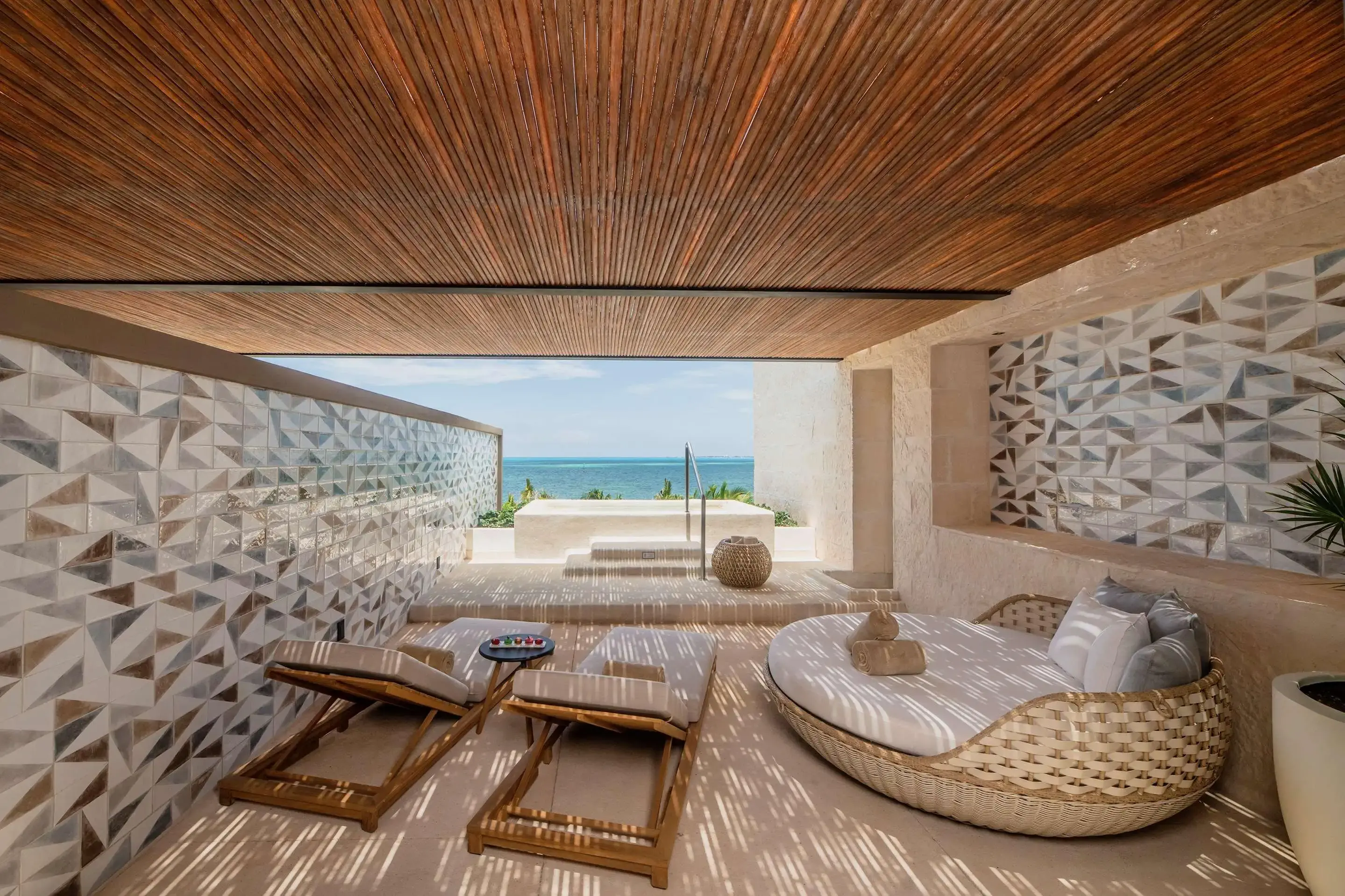 Luxurious beachfront patio with wooden loungers, geometric wall design, and ocean view.