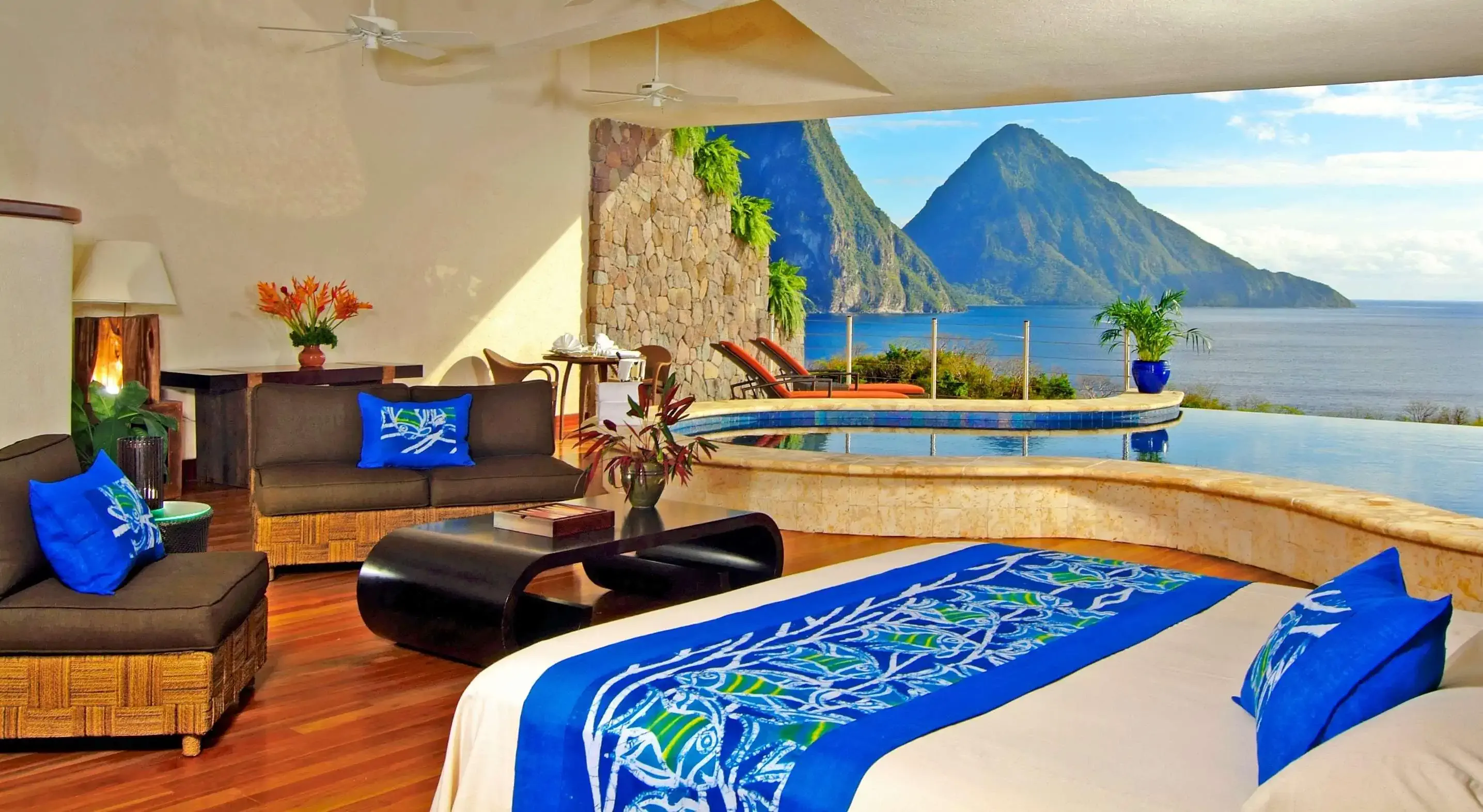 An open-air bedroom with blue tropical linens, an infinity pool, and a stunning view of mountains and the ocean.