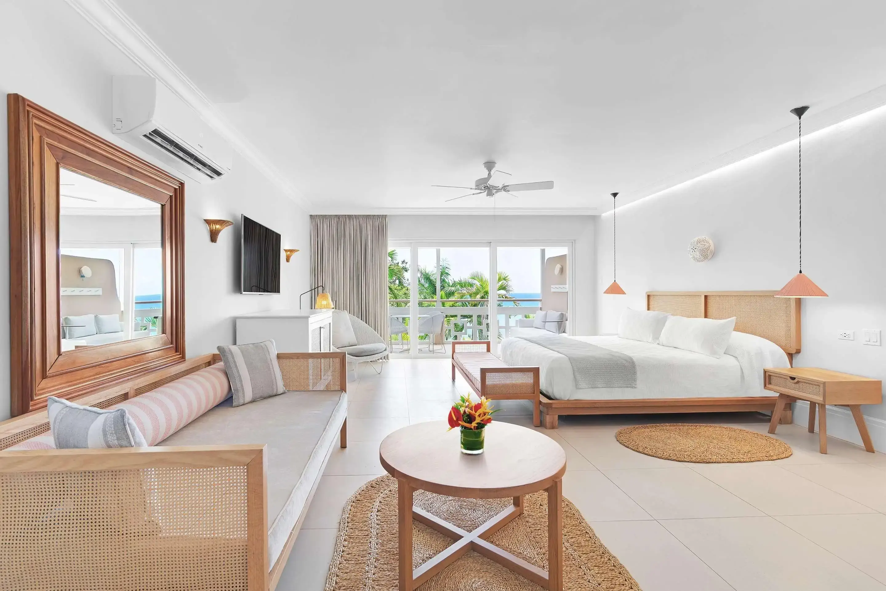 Spacious hotel room with a large bed, sofa, desk, and wall-mounted TV. A glass door leads to a balcony with a view of palm trees