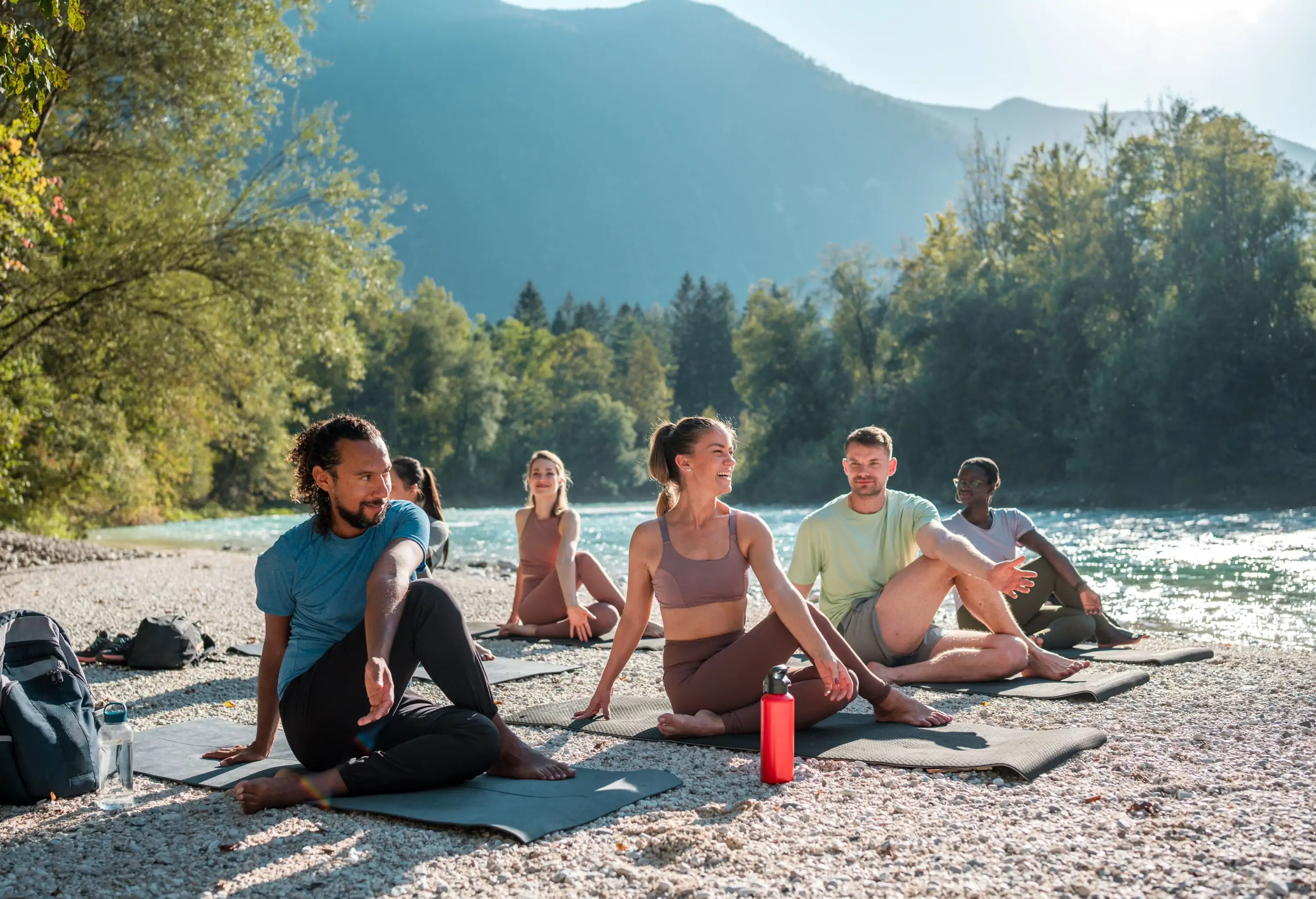 A diverse gathering practices outdoor yoga and stretching on mats by the peaceful riverbank, showcasing a mix of ethnicities.