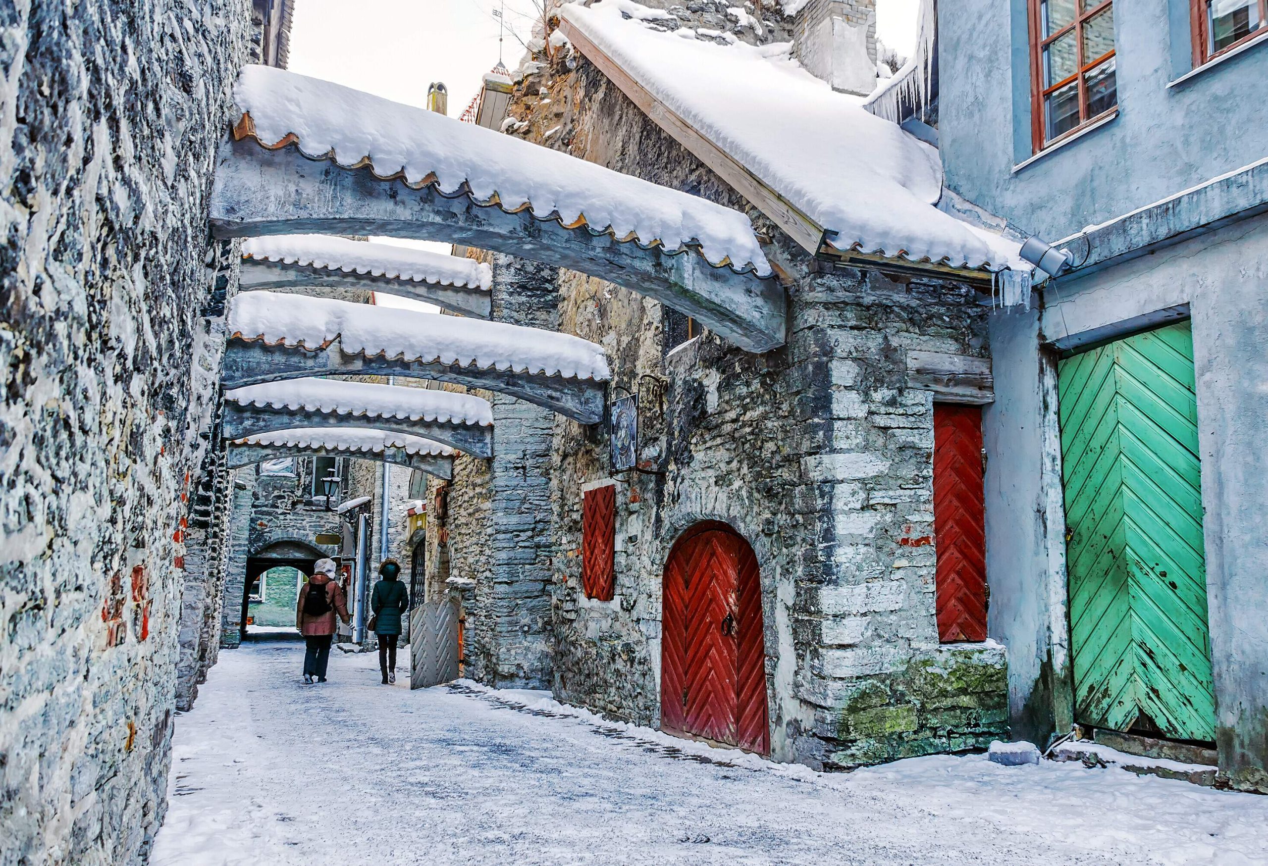Two tourists walk on an alley under the arch ledges of old stone buildings with snow-covered roofs.