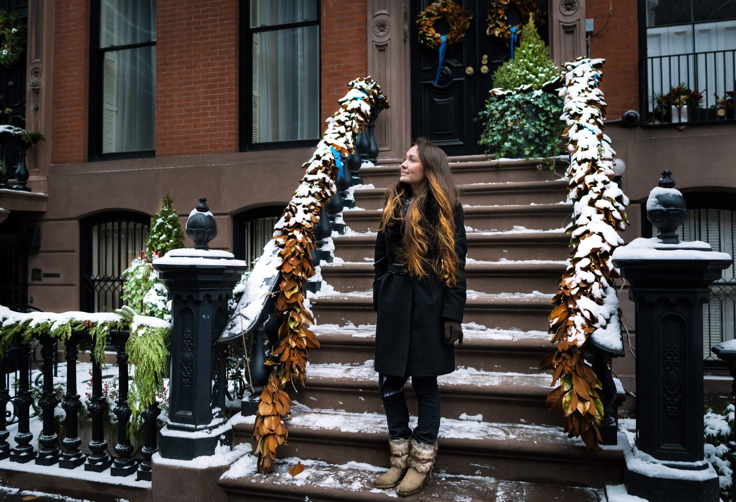 A brunette woman in a black trench coat stands in front of an apartment building's staircase adorned with Christmas decorations.