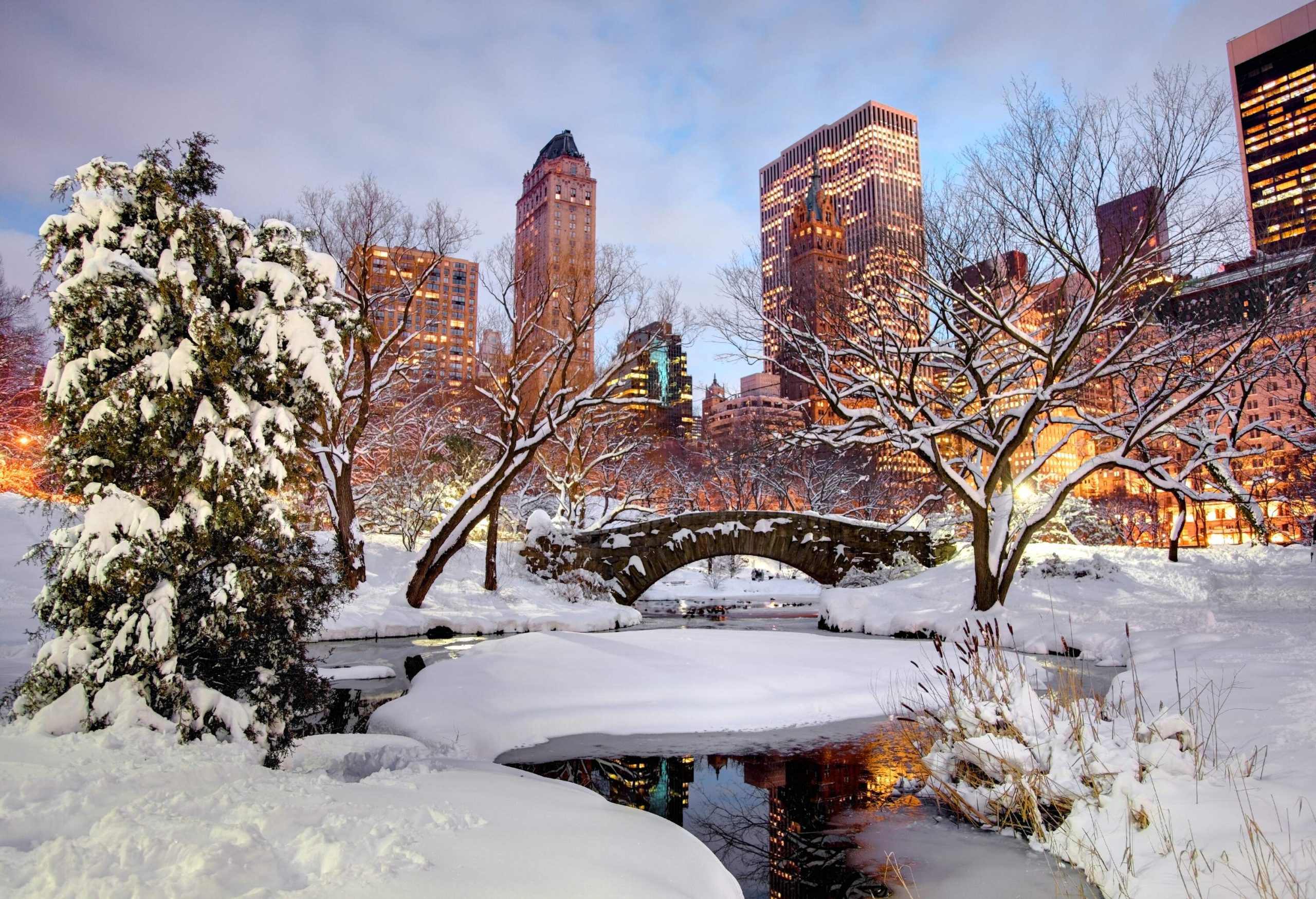 A stone arch bridge across a pond with views of the lit-up skyline and trees covered with snow.