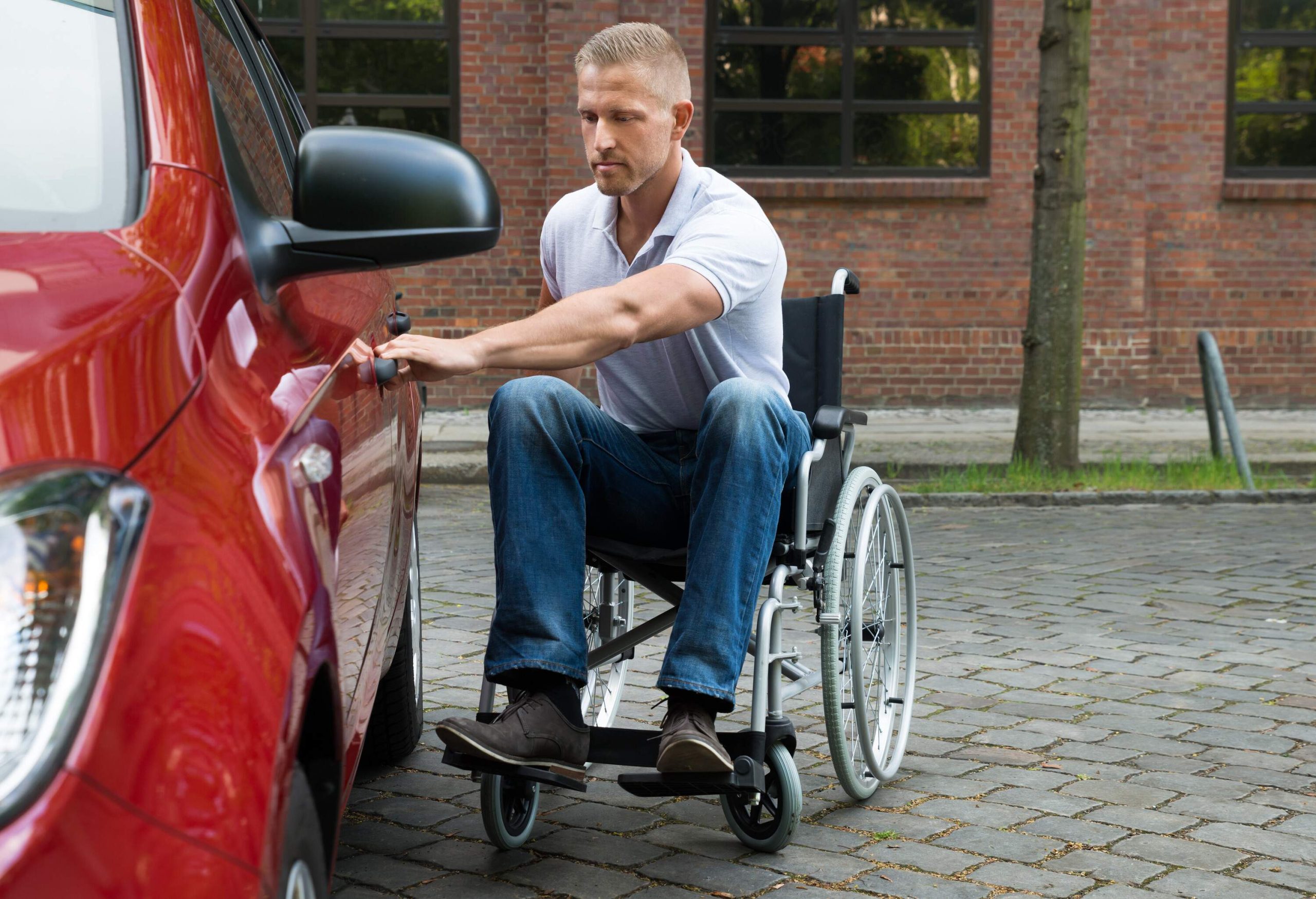 A man sitting in a wheelchair is opening the door of a red car.