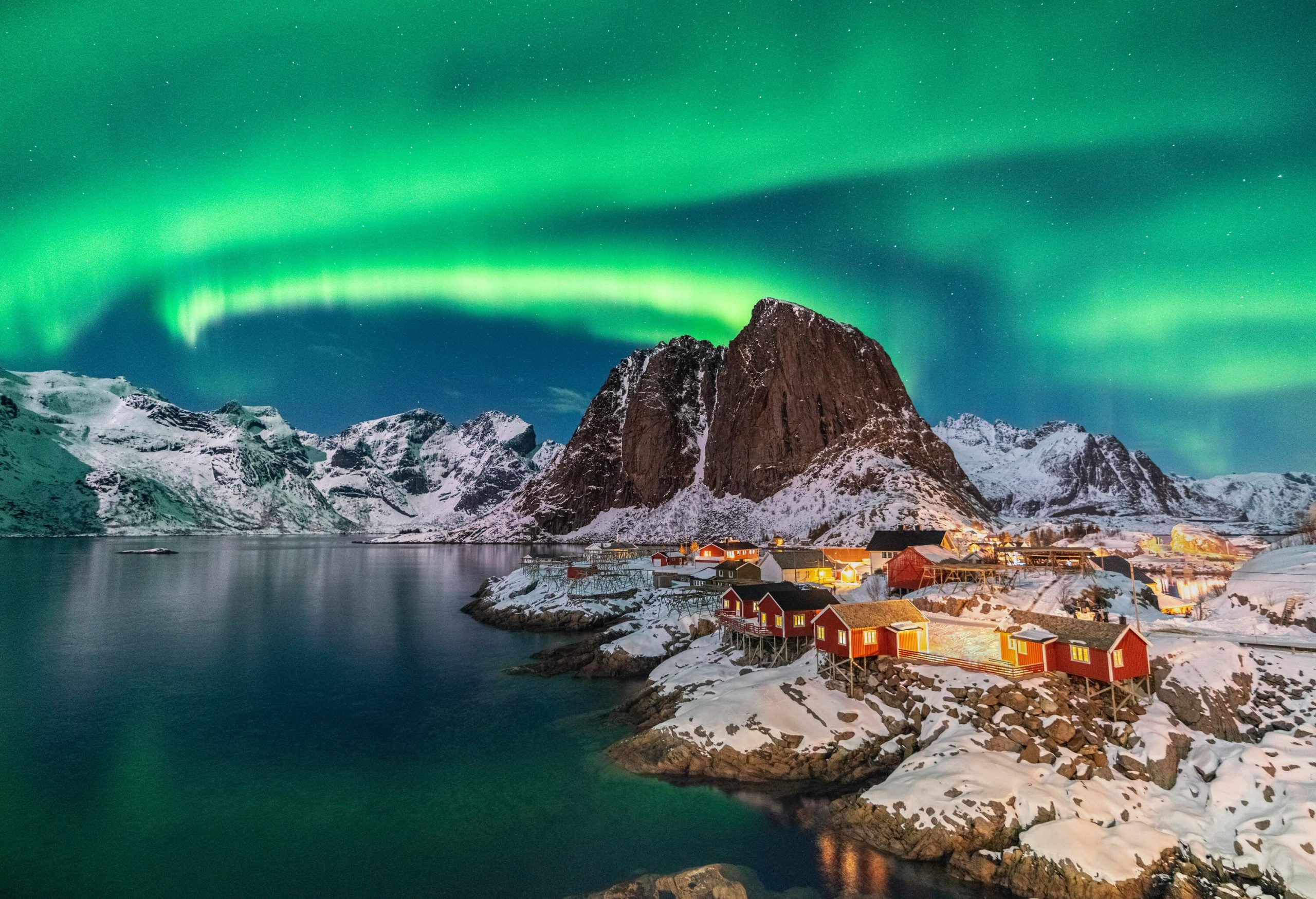 The dazzling Northern lights colour the sky green and blue over a small village along the lake bounded by steep rocks.