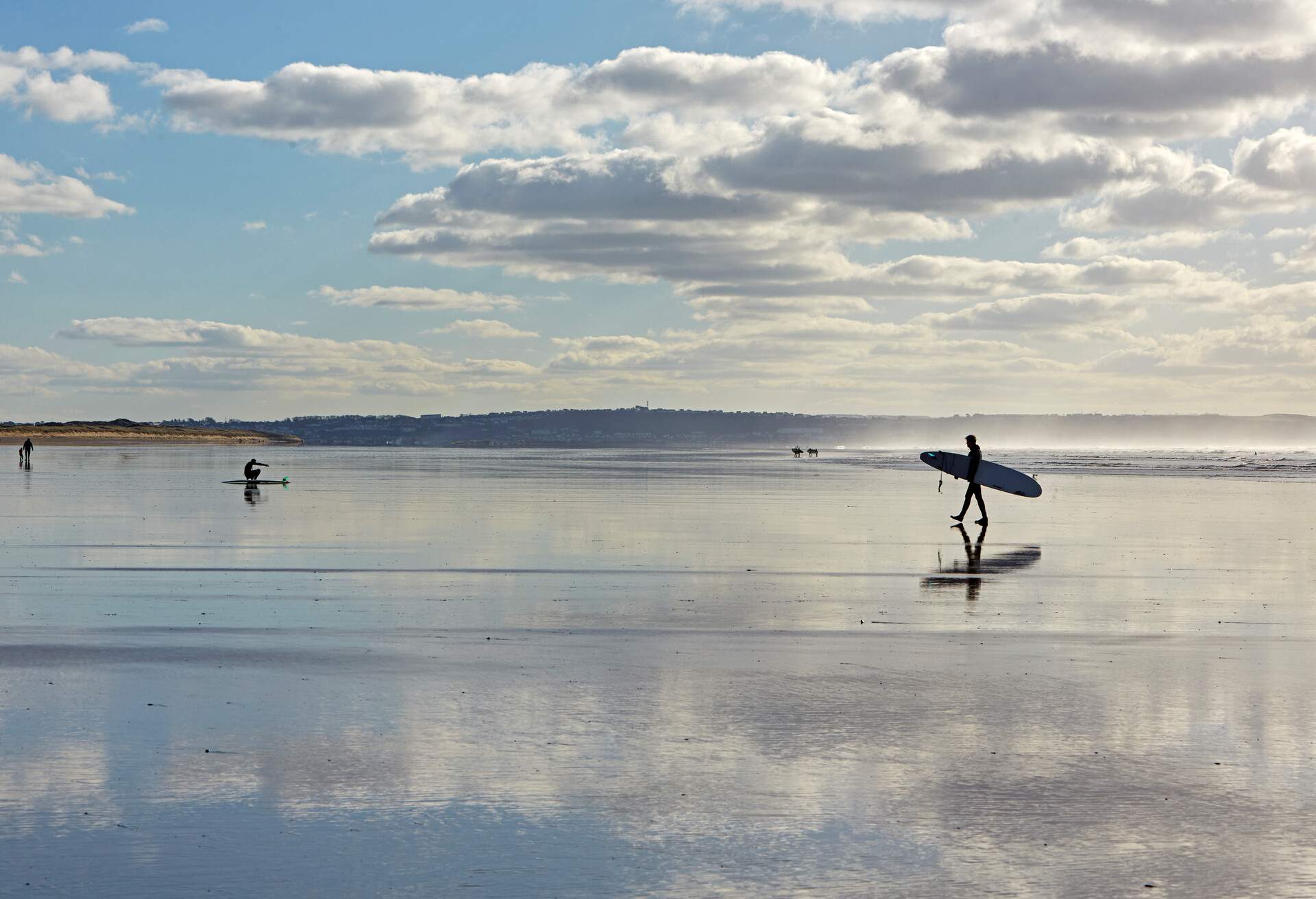 Silhouette of man sitting next to surfboard and surfer across Saunton Sands beach in North Devon at low tide