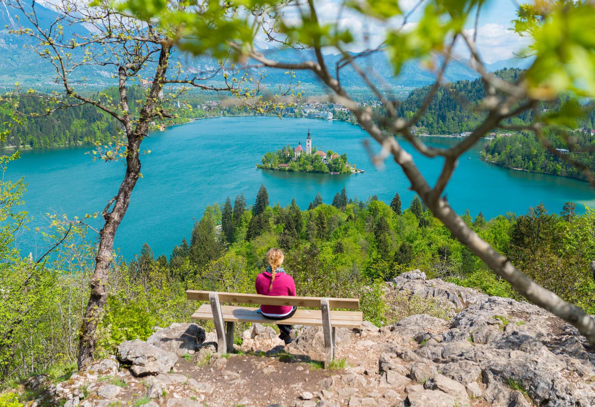 A person sits on a hilltop bench and looks at the church island in the middle of a lake.