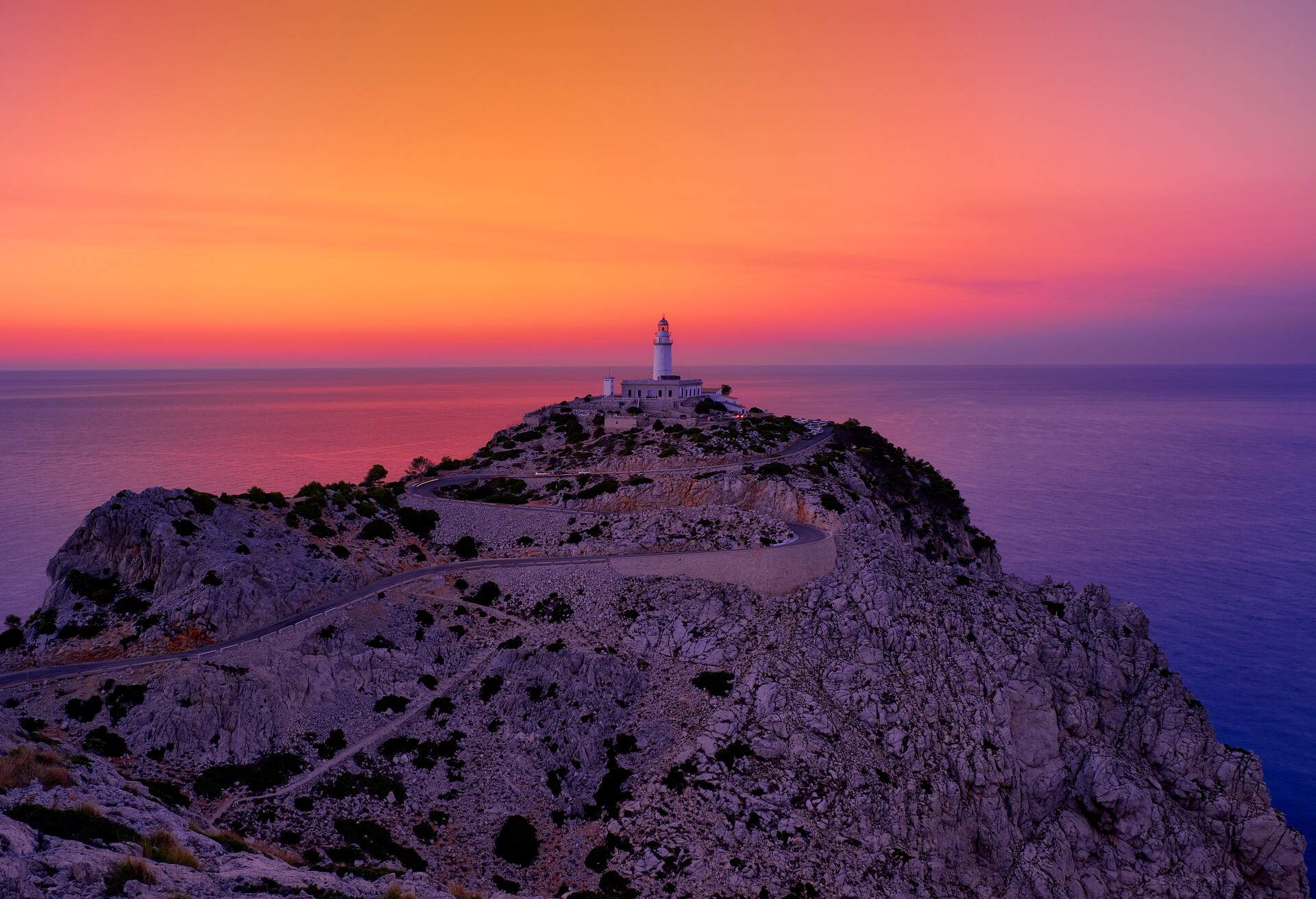 A lighthouse on top of a rocky mountain by the sea with a dramatic purple-orange sky.