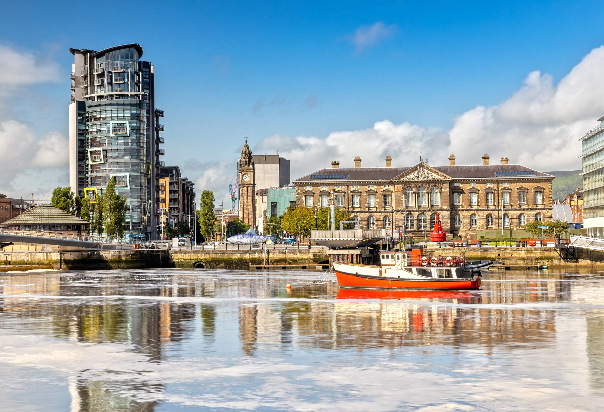 A fishing boat anchored on a river, with harbourfront historic buildings and a clock tower.