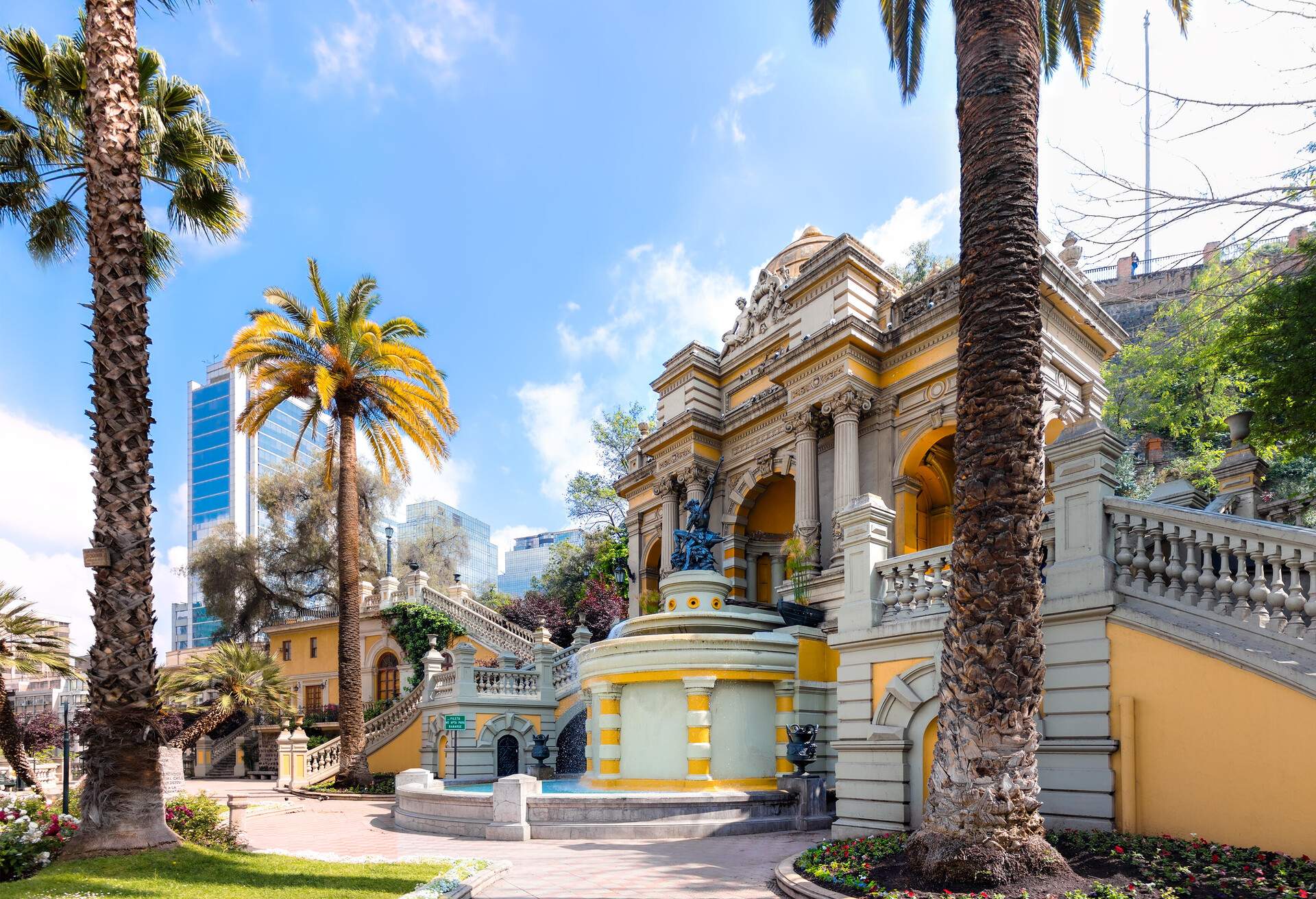 A fountain with a sculpture atop a cylindrical pedestal in front of a colonial edifice.