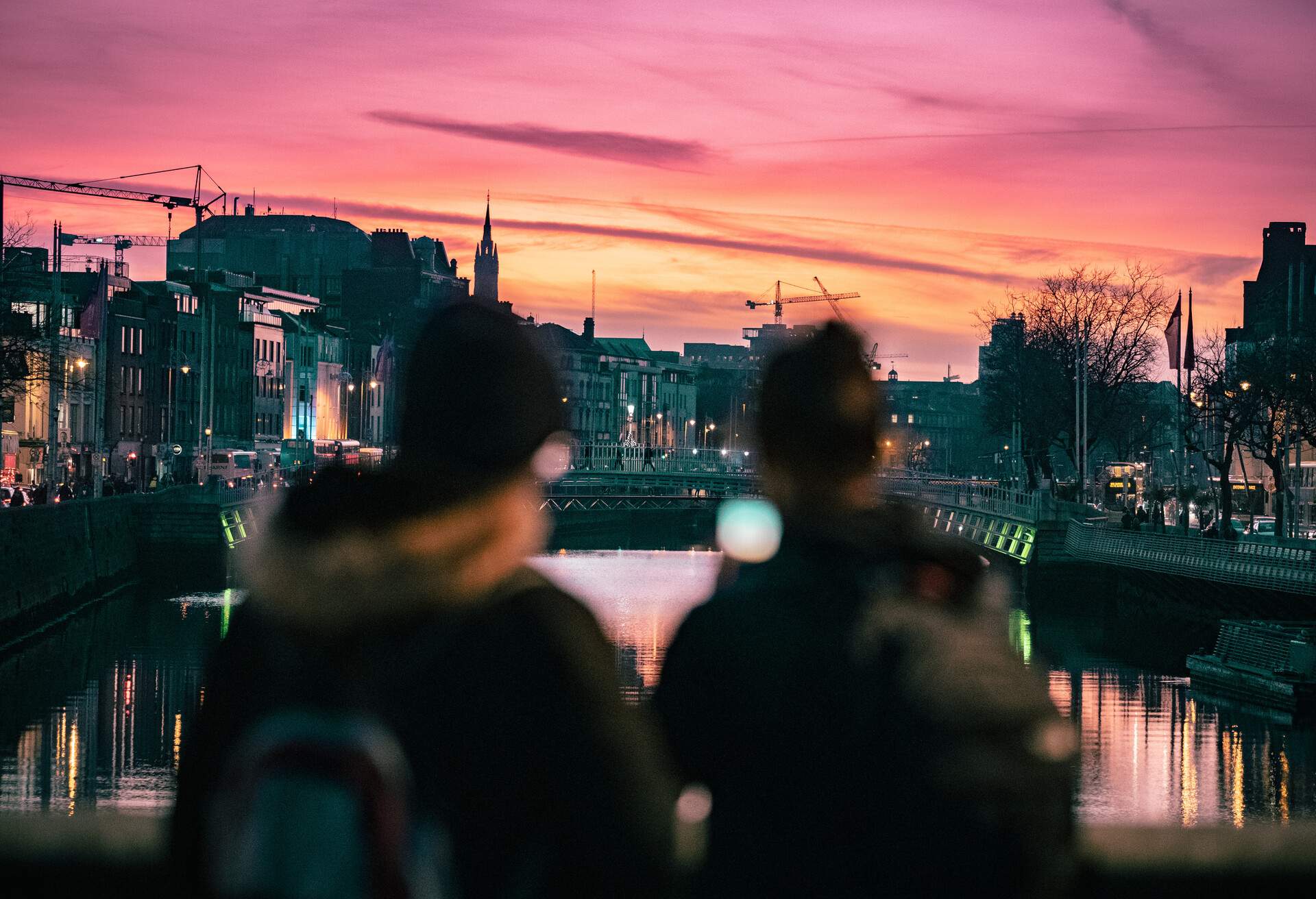 Against the backdrop of the city skyline, two vague figures can be seen standing on a bridge above the river.