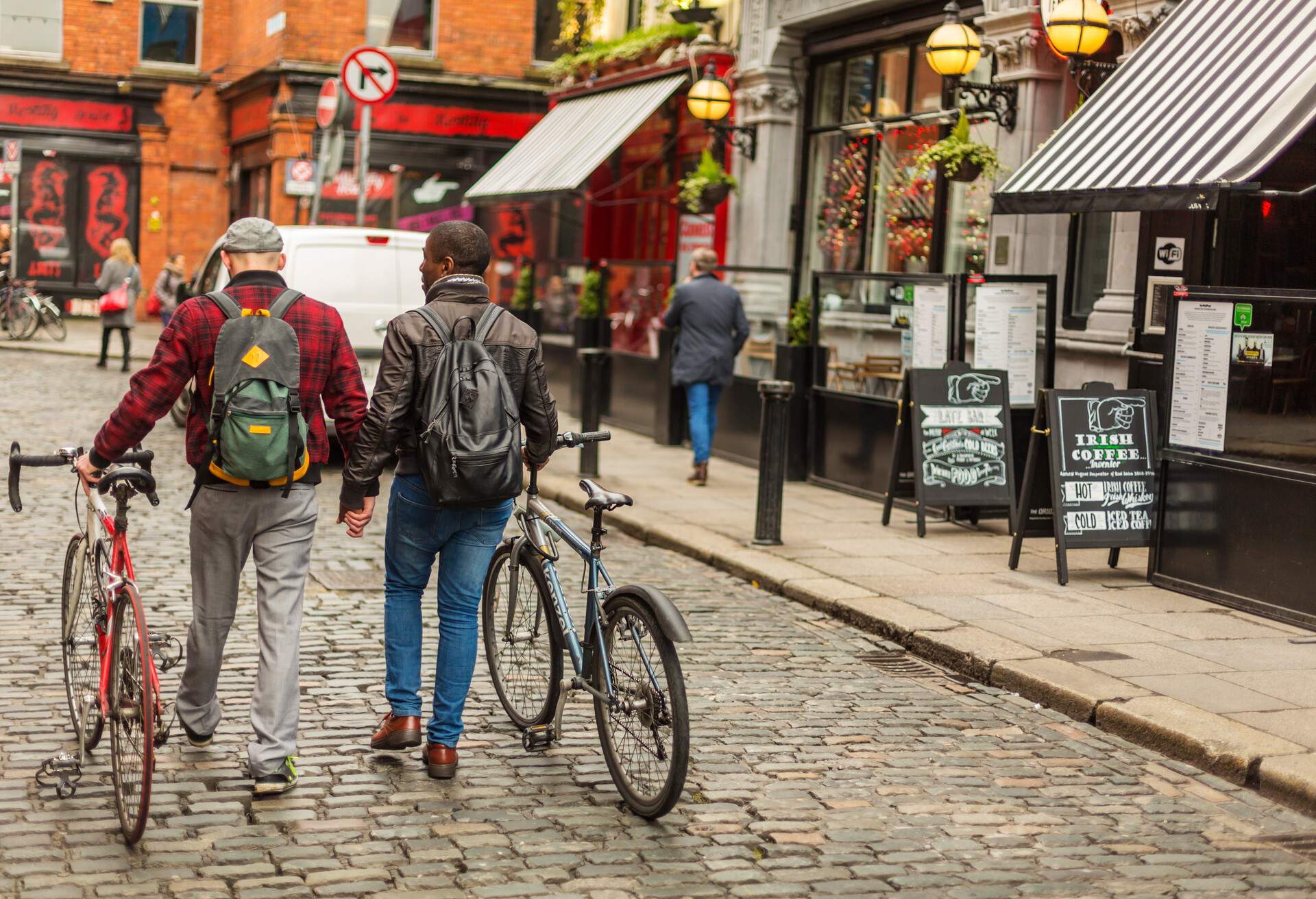 A gay couple in winter clothes and backpacks hold hands as they tow their bikes on the cobblestone street in front of the stores.