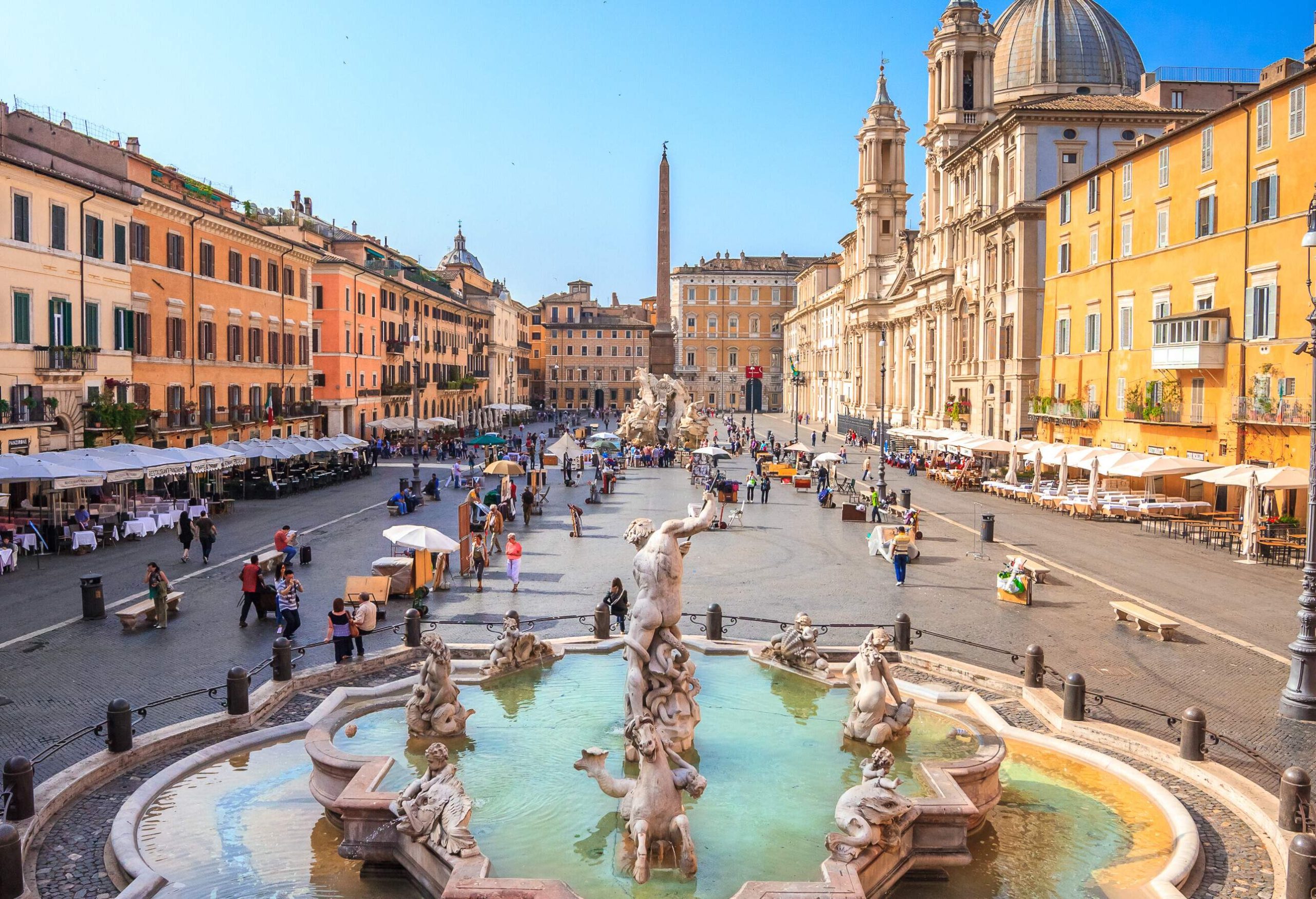 The Fontana del Nettuno, a magnificent Baroque fountain featuring a statue of Neptune surrounded by sea creatures and cherubs, is located in a bustling square with shops, cafes, and historic buildings.
