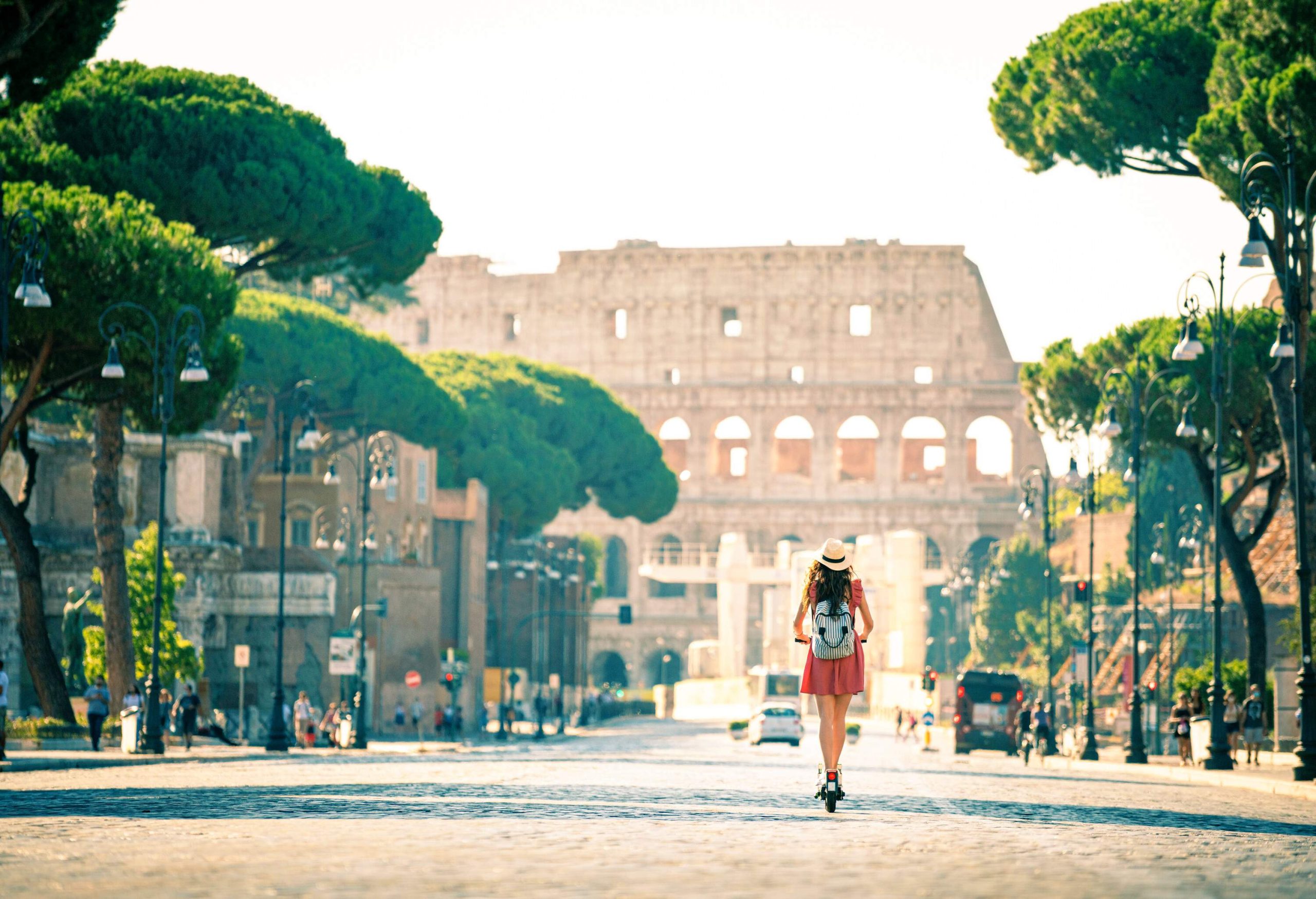 A woman in casual clothes rides an electric scooter across a tree-lined cobblestone street towards the Colosseum.