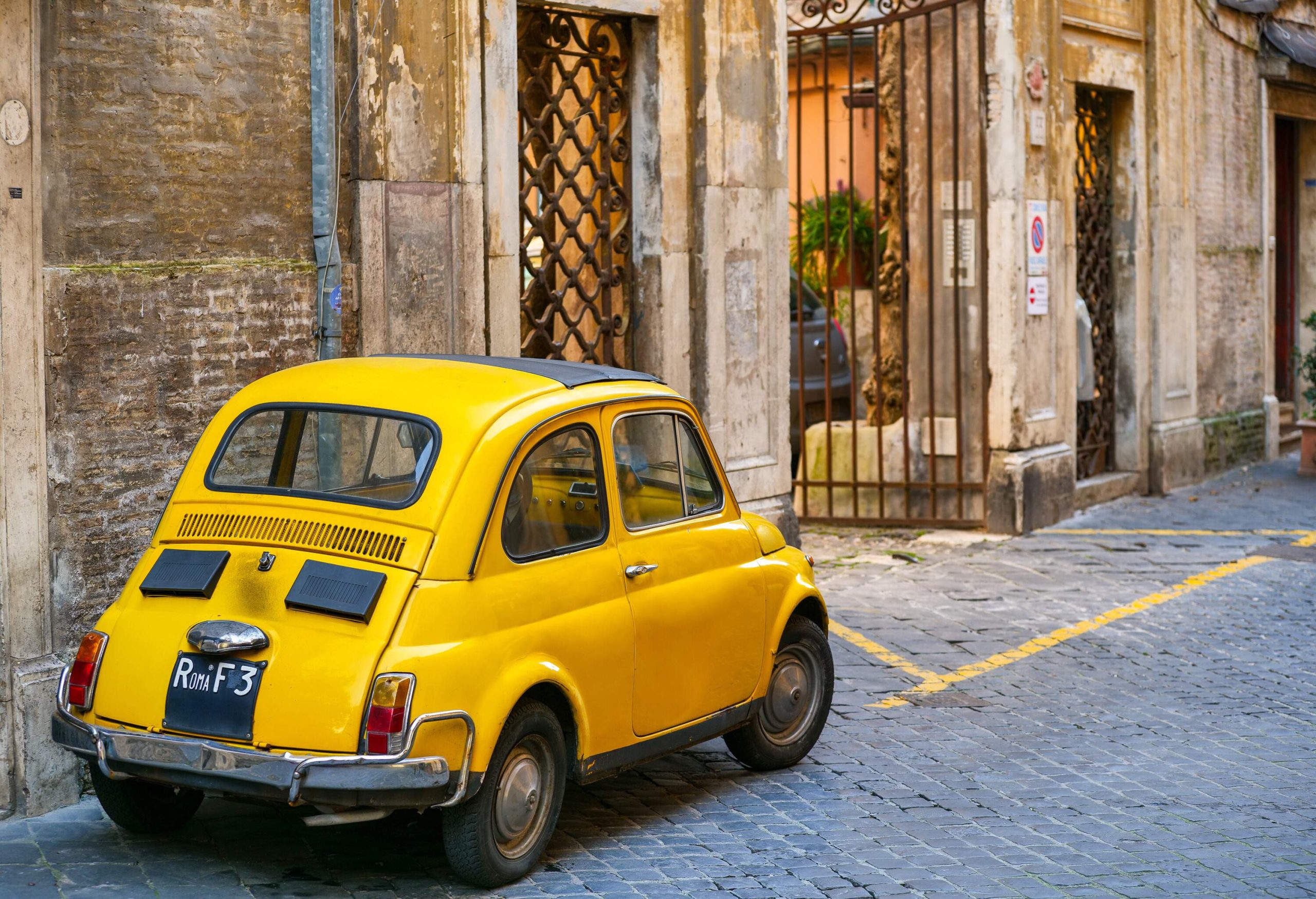 A yellow vintage car parked beside a gate along an alley.