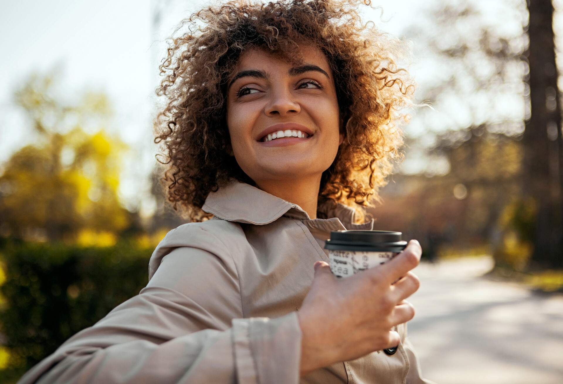 A smiling curly-hair woman sitting on a park bench with a to go coffee during fall