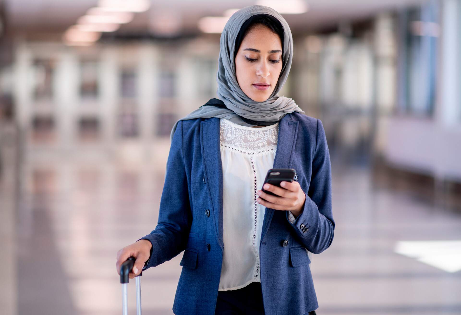 Happy solo female traveler texts on a mobile phone while waiting inside the airport with luggage.